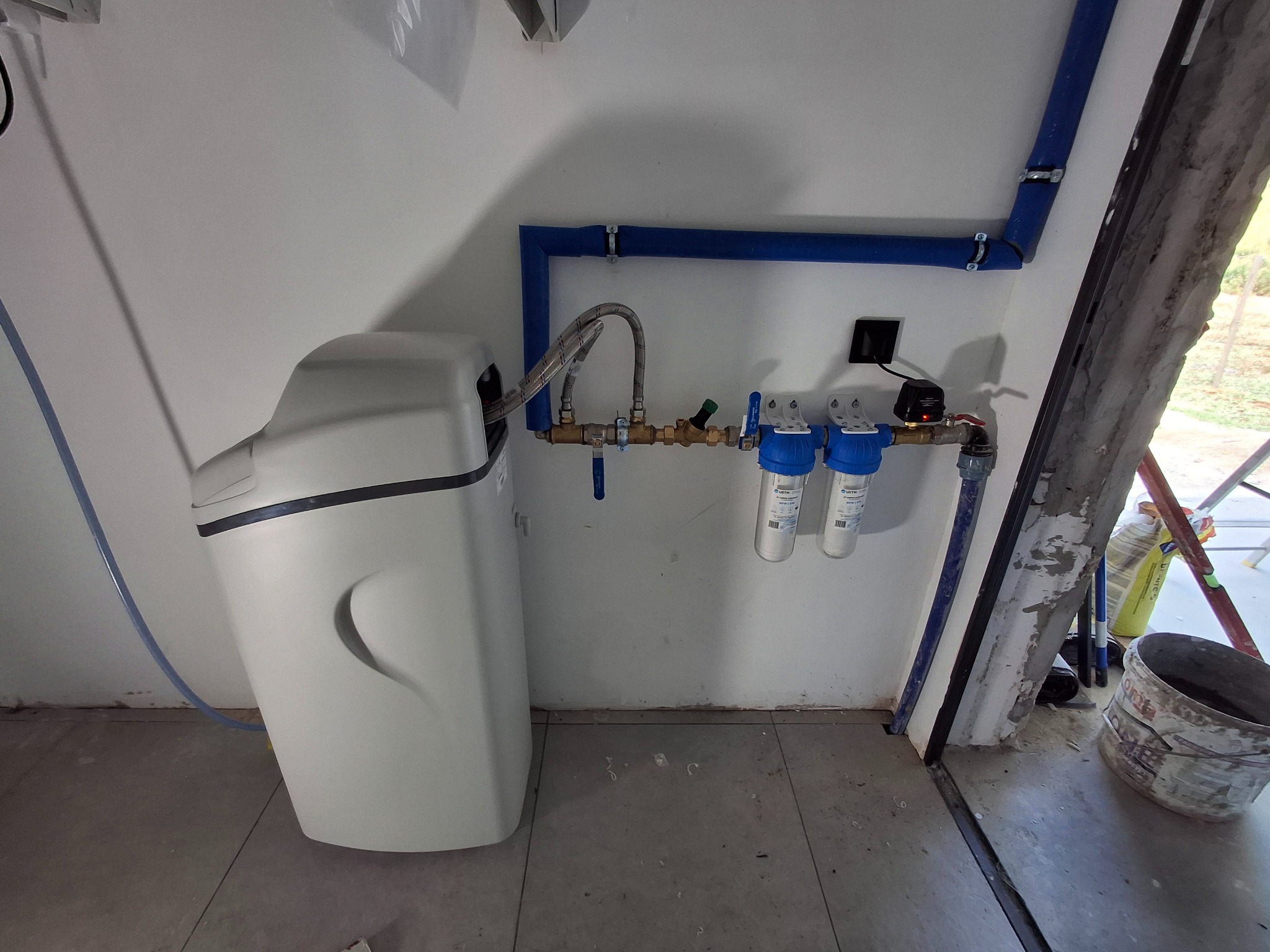 Biały zmiękczacz wody i filtry zamontowane na ścianie z niebieskimi rurami. Widoczne także elementy instalacji i wiadro. Ujęcie z bliska, w pomieszczeniu.