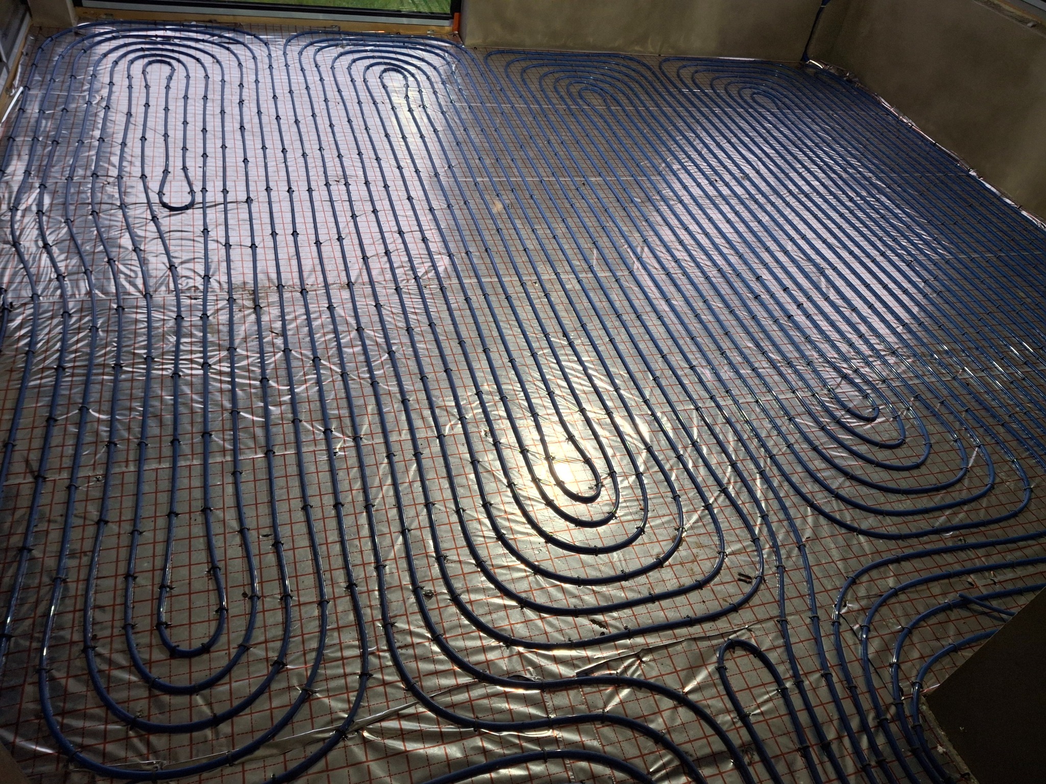 Niebieskie rury ogrzewania podłogowego ułożone na folii z siatką, widok z góry. Instalacja w trakcie realizacji, równoległe ułożenie z zakręconymi zakończeniami.