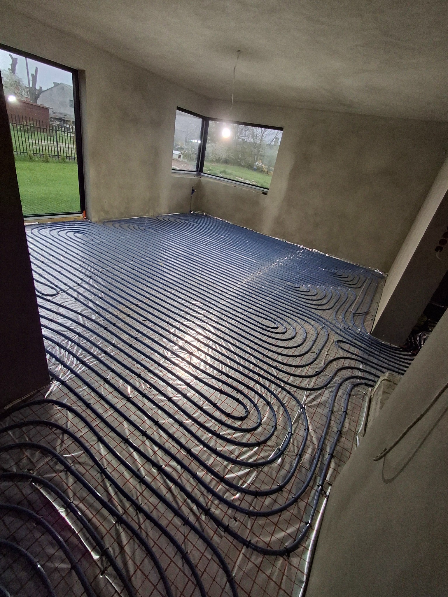 Niebieskie rury ogrzewania podłogowego ułożone na folii z siatką, widoczne okna i surowe ściany w pomieszczeniu przed wylewką.