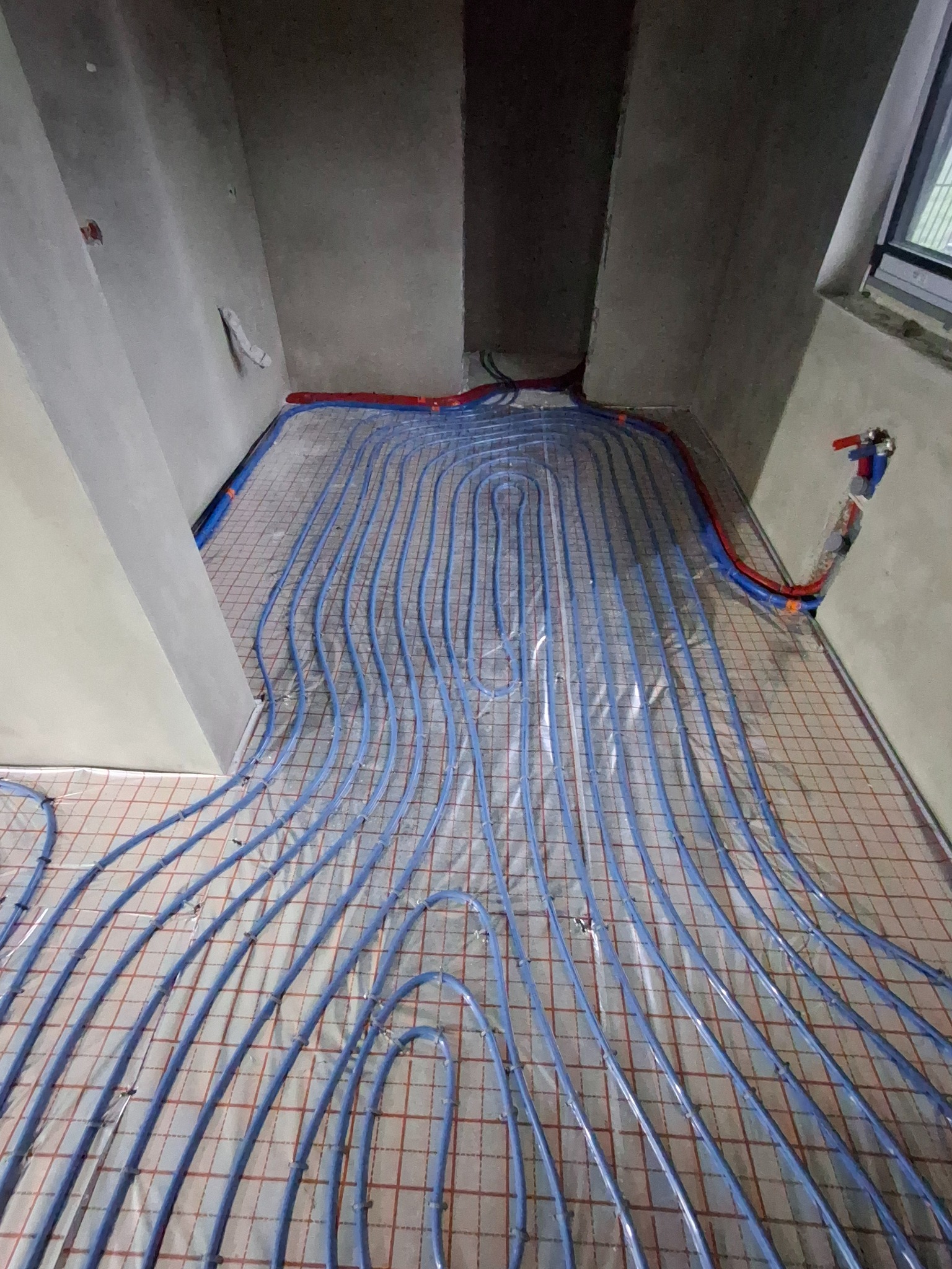 Niebieskie rury ogrzewania podłogowego ułożone na izolacji z siatką w surowym wnętrzu. Widoczne przyłącza i fragment okna.