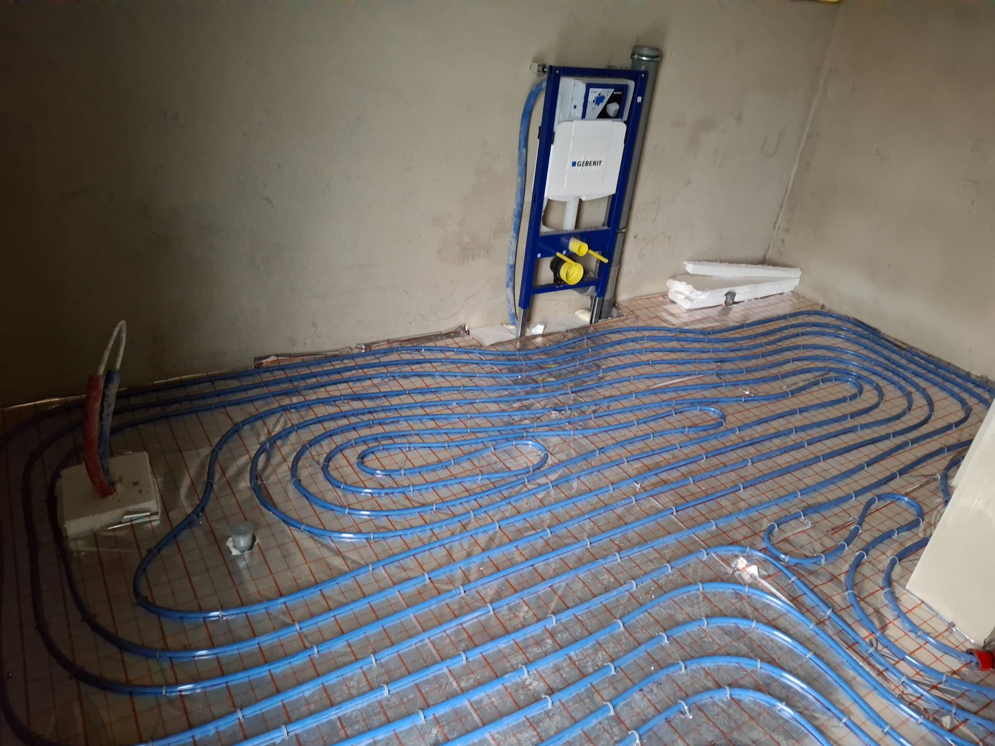 Niebieskie rury ogrzewania podłogowego ułożone na siatce grzewczej w surowym wnętrzu, z widocznym stelażem podtynkowym Geberit i elementami instalacji.