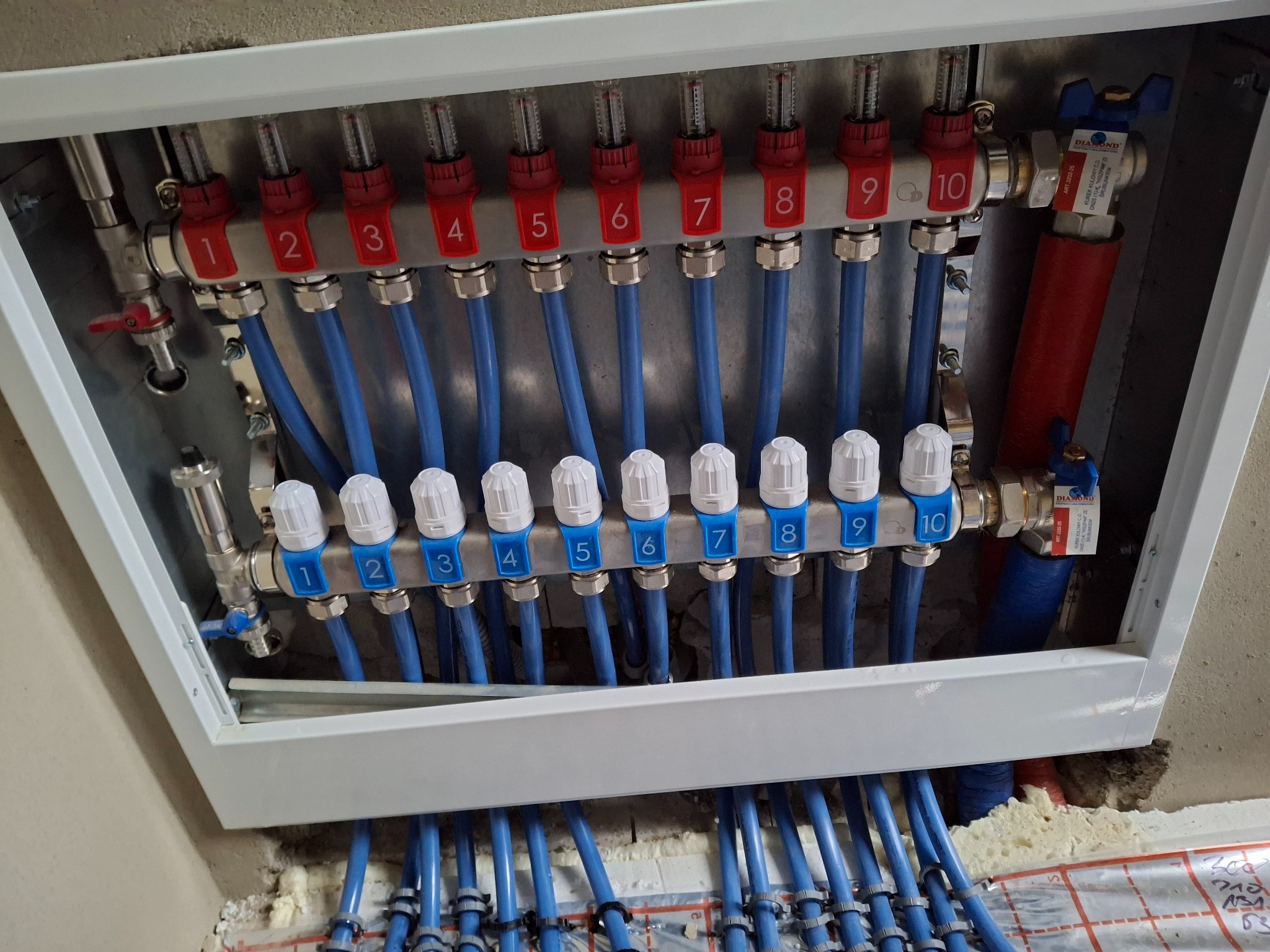 Rozdzielacz ogrzewania podłogowego z dziesięcioma obwodami, z numeracją od 1 do 10, w białej obudowie, z widocznymi niebieskimi rurami i elementami regulacyjnymi.