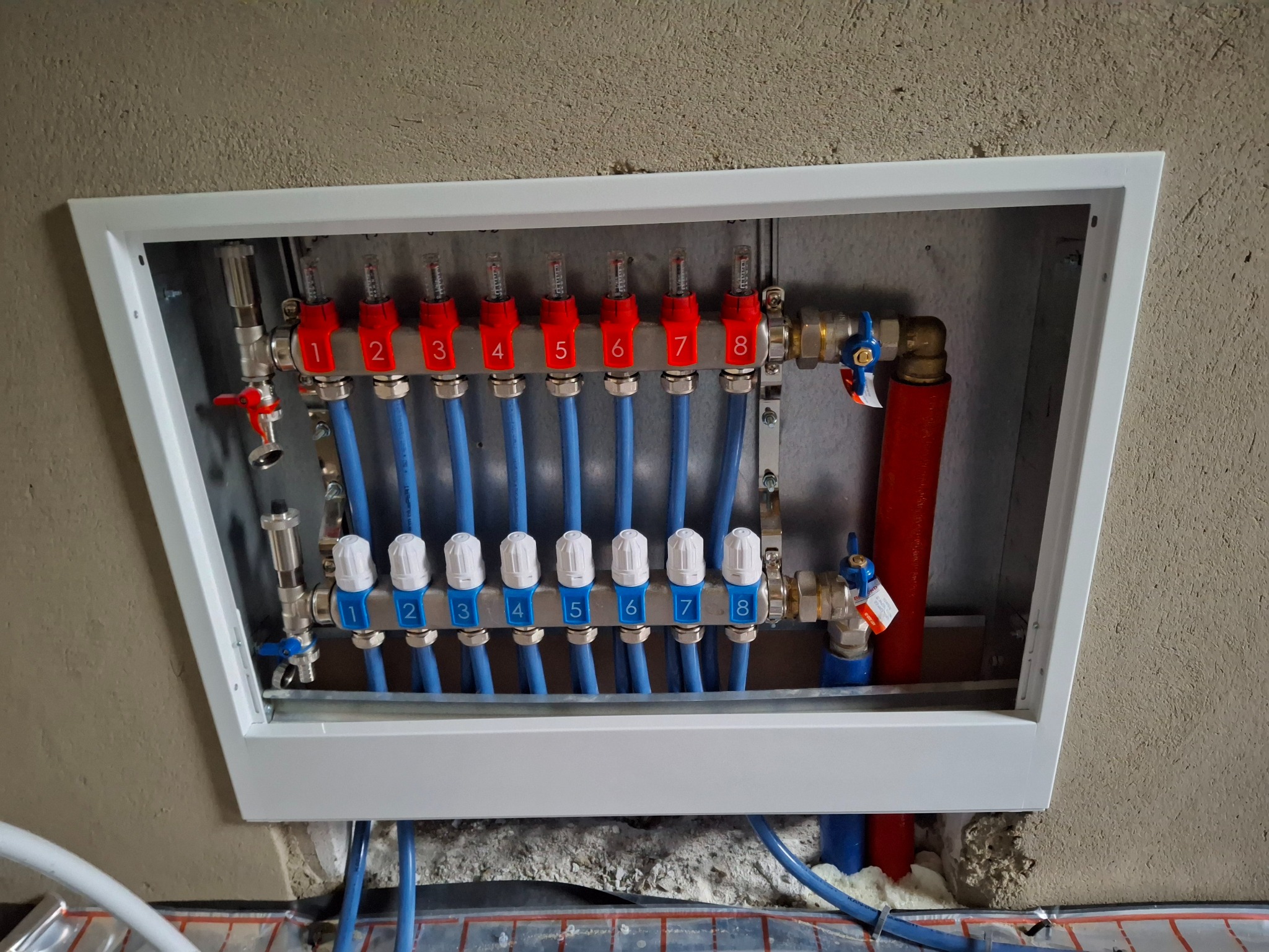 Rozdzielacz ogrzewania podłogowego w białej obudowie, z numerowanymi sekcjami (1-8), niebieskimi rurami i czerwonymi elementami regulacyjnymi, zamontowany na surowej ścianie.