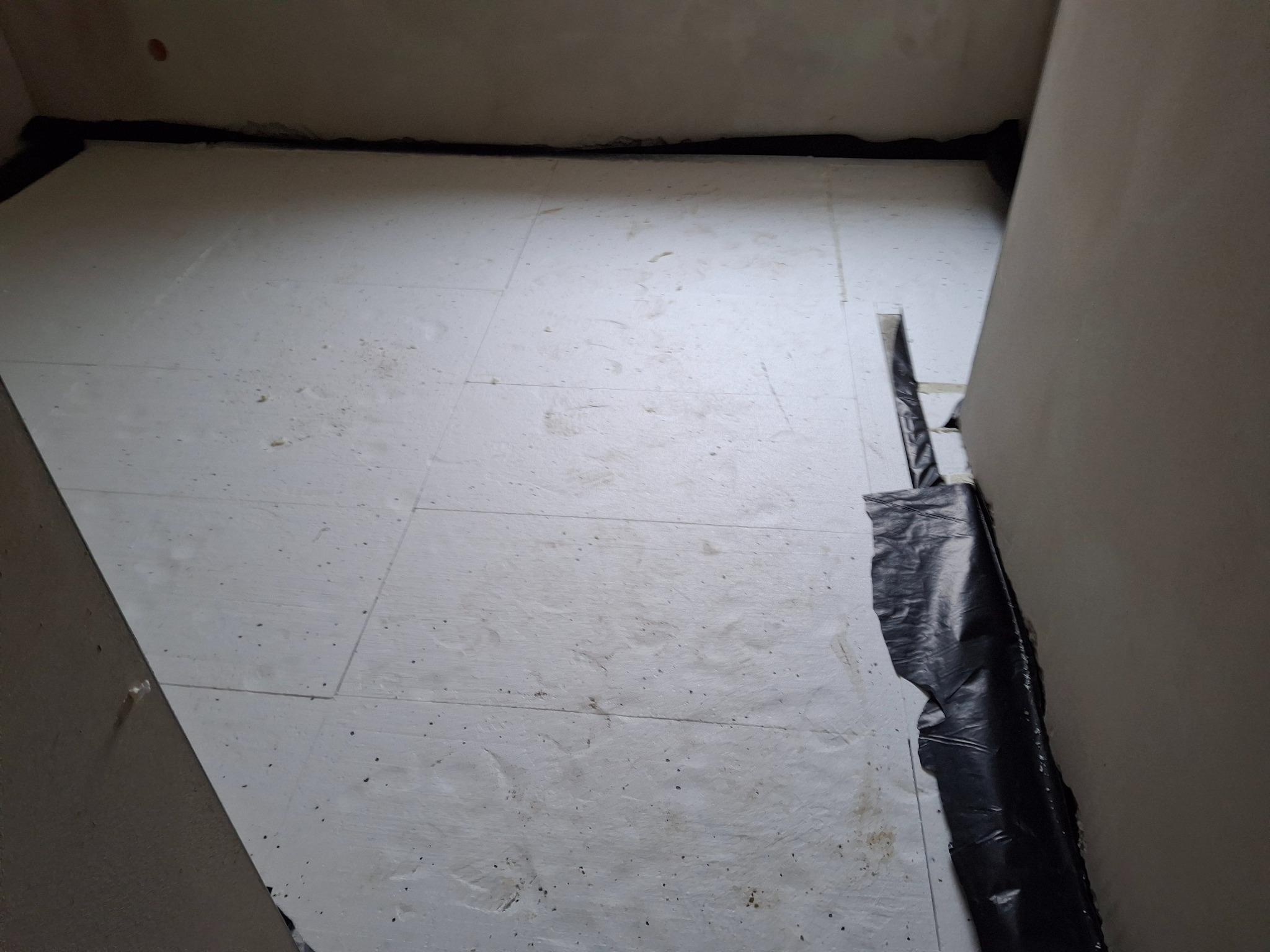 Białe płyty izolacyjne pod ogrzewanie podłogowe, zabezpieczone czarną folią przy ścianach. Przygotowanie do wylewki, widok z góry, surowy stan.