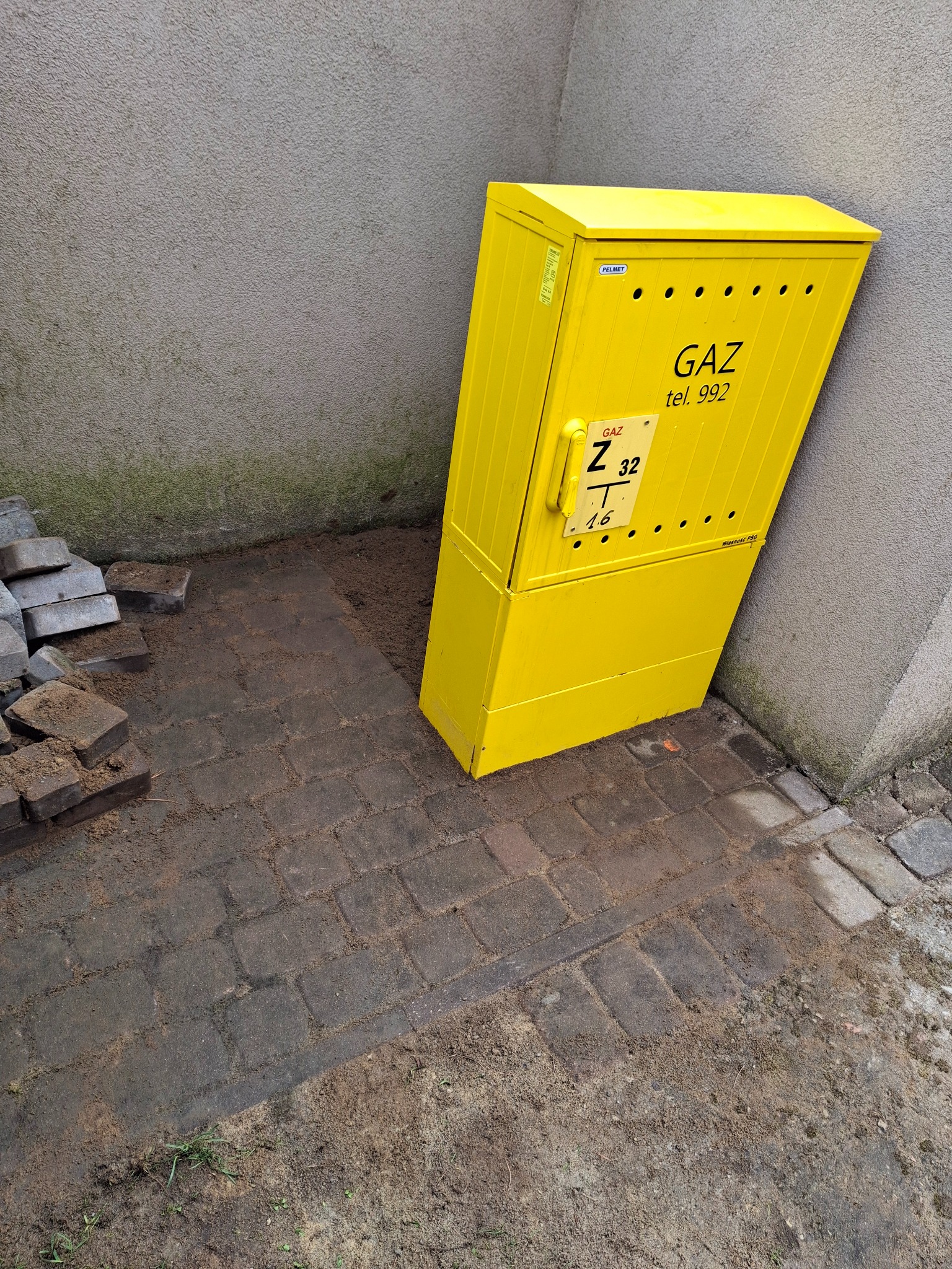 Żółta skrzynka gazowa przy szarej ścianie, z widocznymi cegłami i ziemią wokół. Skrzynka oznaczona napisem 'GAZ tel. 992' i numerem Z 32 T 16.