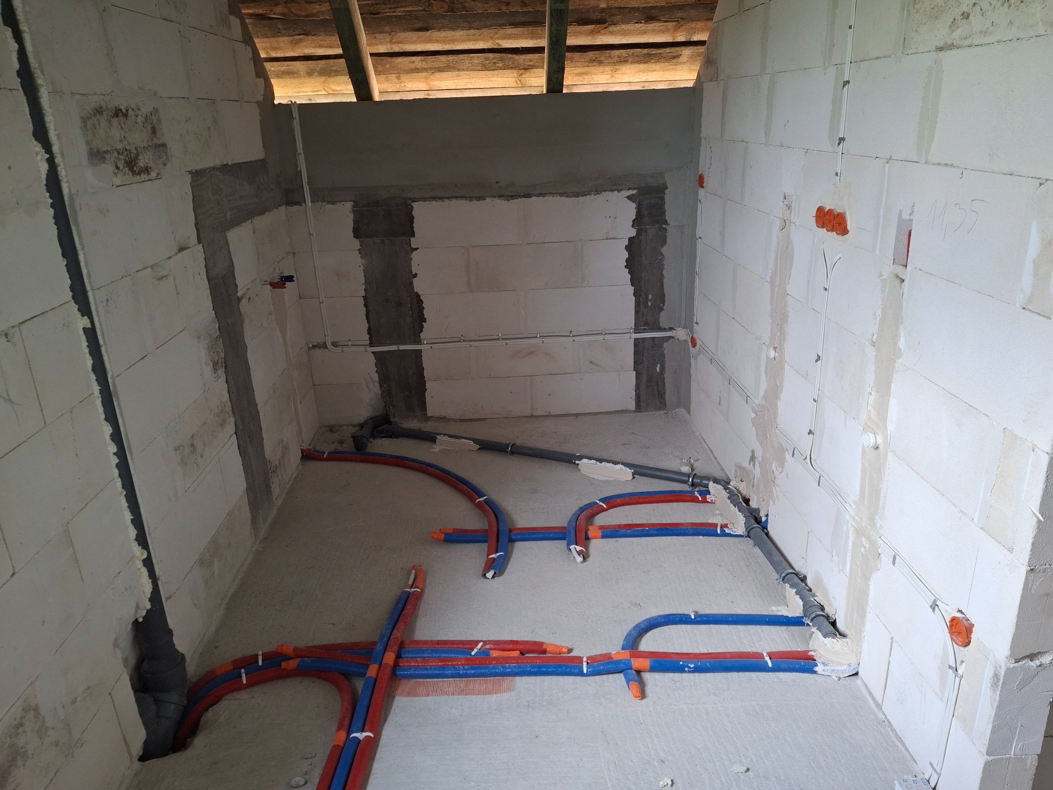 Wnętrze w trakcie instalacji: widoczne rury CO (czerwono-niebieskie) na podłodze i peszle elektryczne w ścianach z betonu komórkowego. Surowy stan, instalacje w toku.
