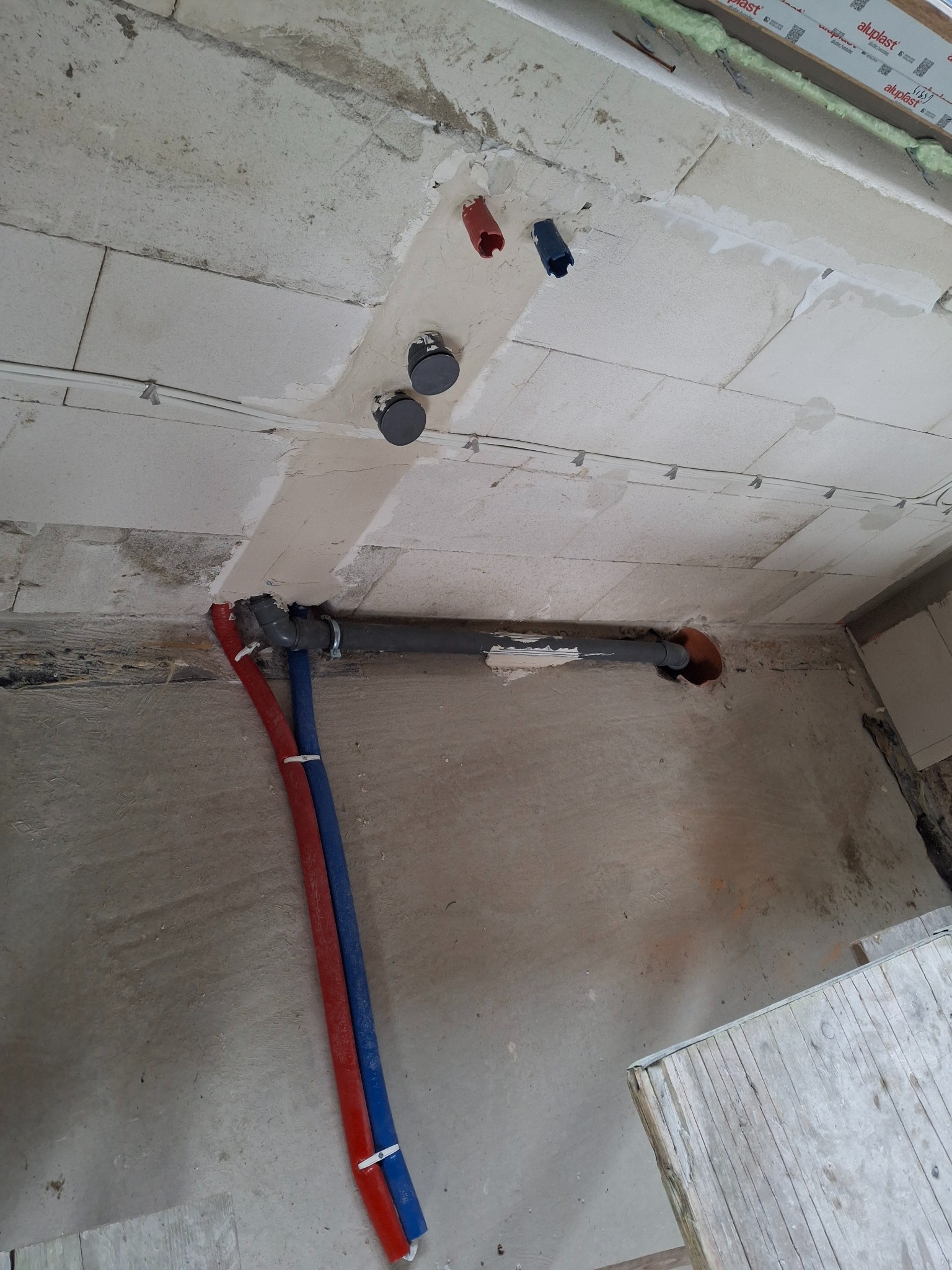 Instalacja rur kanalizacyjnych i wodnych w surowym wnętrzu. Widoczne rury w kolorach czerwonym i niebieskim, prowadzone wzdłuż ściany, oraz szara rura kanalizacyjna.