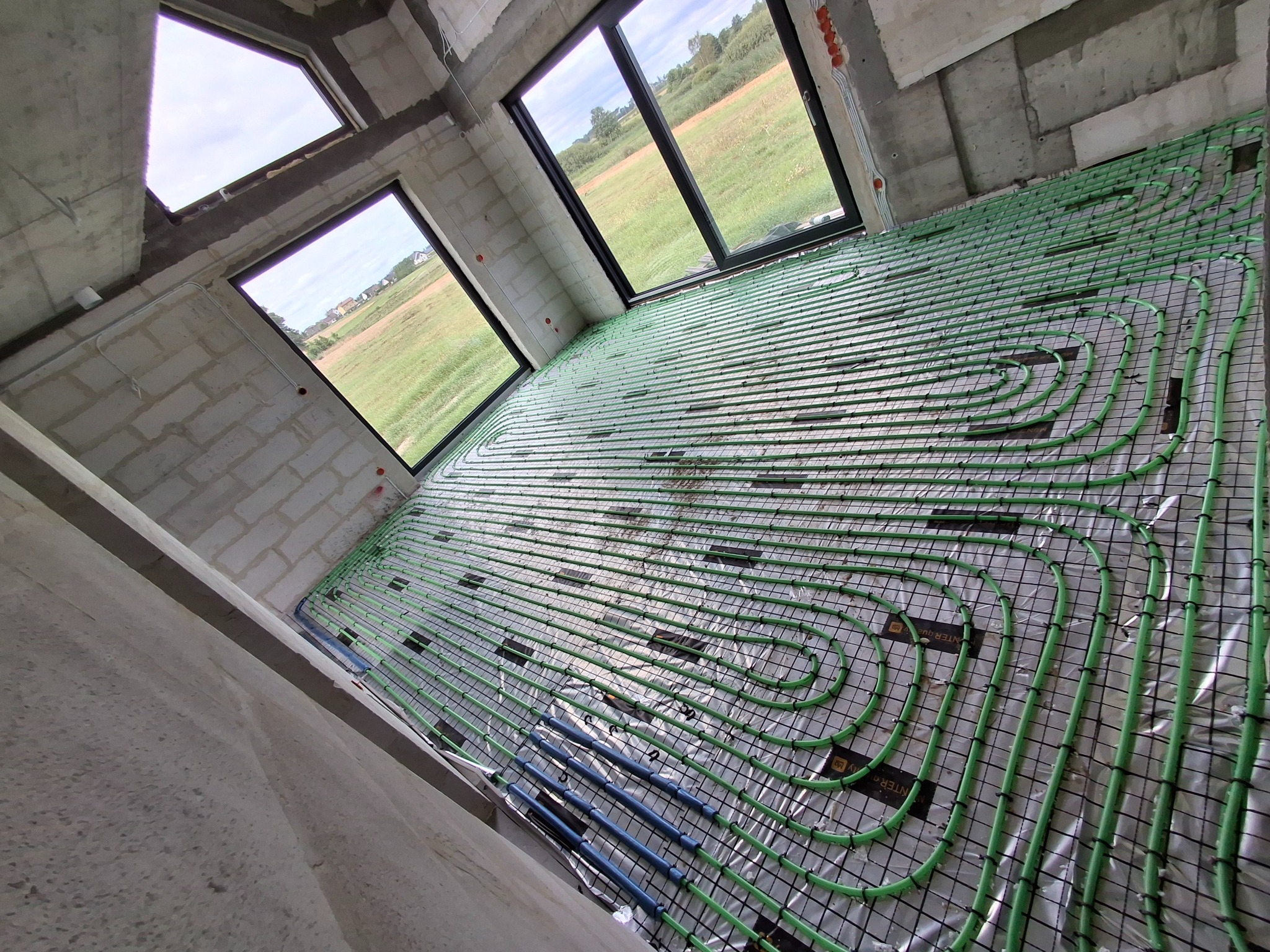 Ogrzewanie podłogowe w surowym wnętrzu w Rąbiu: zielone rurki na czarnej siatce, widok z góry, okna z widokiem na zewnątrz, surowy beton.