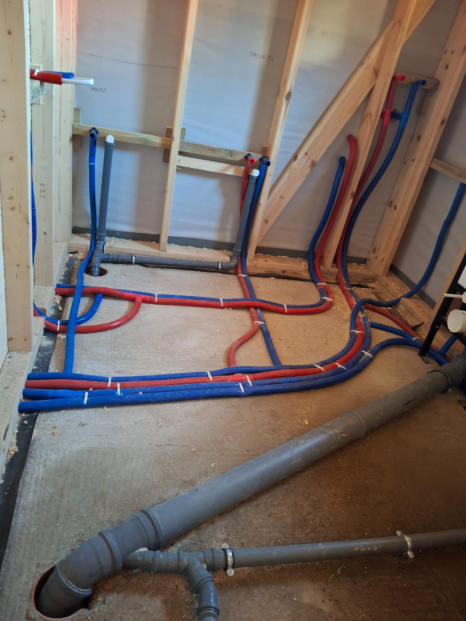 Instalacja wodna i kanalizacyjna w surowym wnętrzu poddasza. Czerwone i niebieskie rury prowadzone po podłodze i ścianach, szare rury kanalizacyjne w dolnej części kadru.