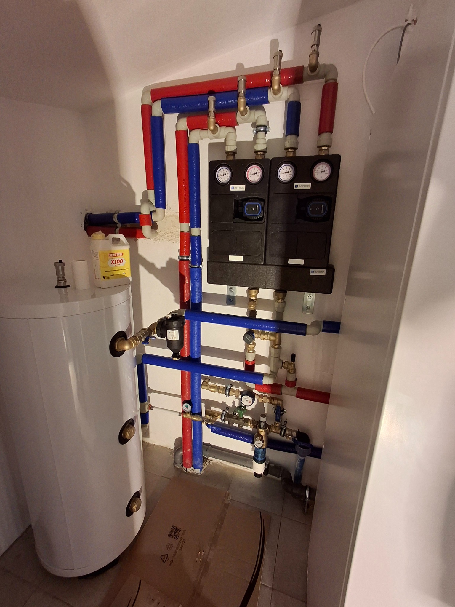 Węzeł cieplny z zasobnikiem CWU, rury w izolacji czerwonej i niebieskiej, manometry na rozdzielaczu, białe ściany, widok z boku, instalacja w pomieszczeniu technicznym.