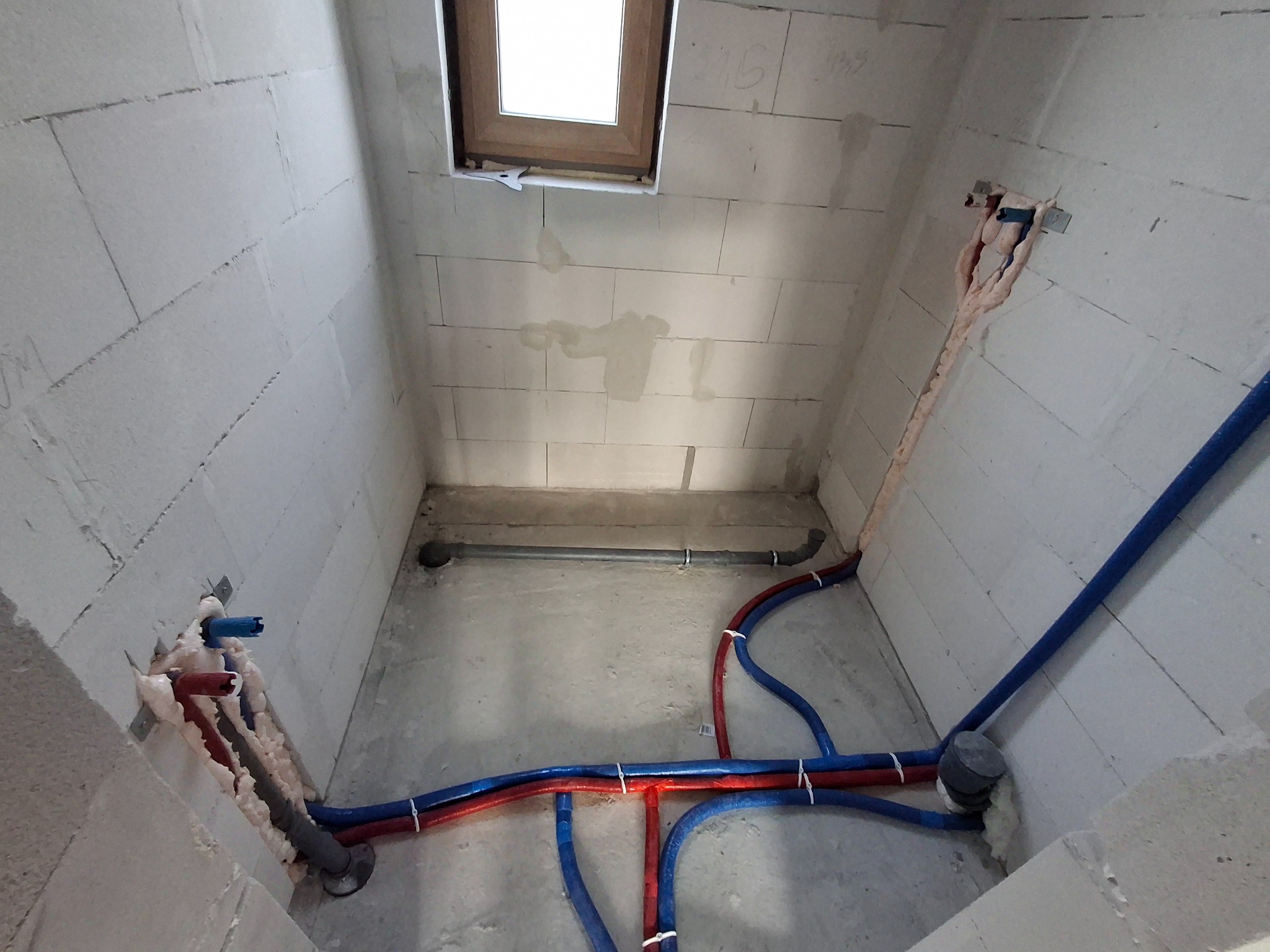 Surowy stan łazienki z widoczną instalacją hydrauliczną: rury PEX w izolacji, odpływ kanalizacyjny i okno dachowe. Ściany z betonu komórkowego.