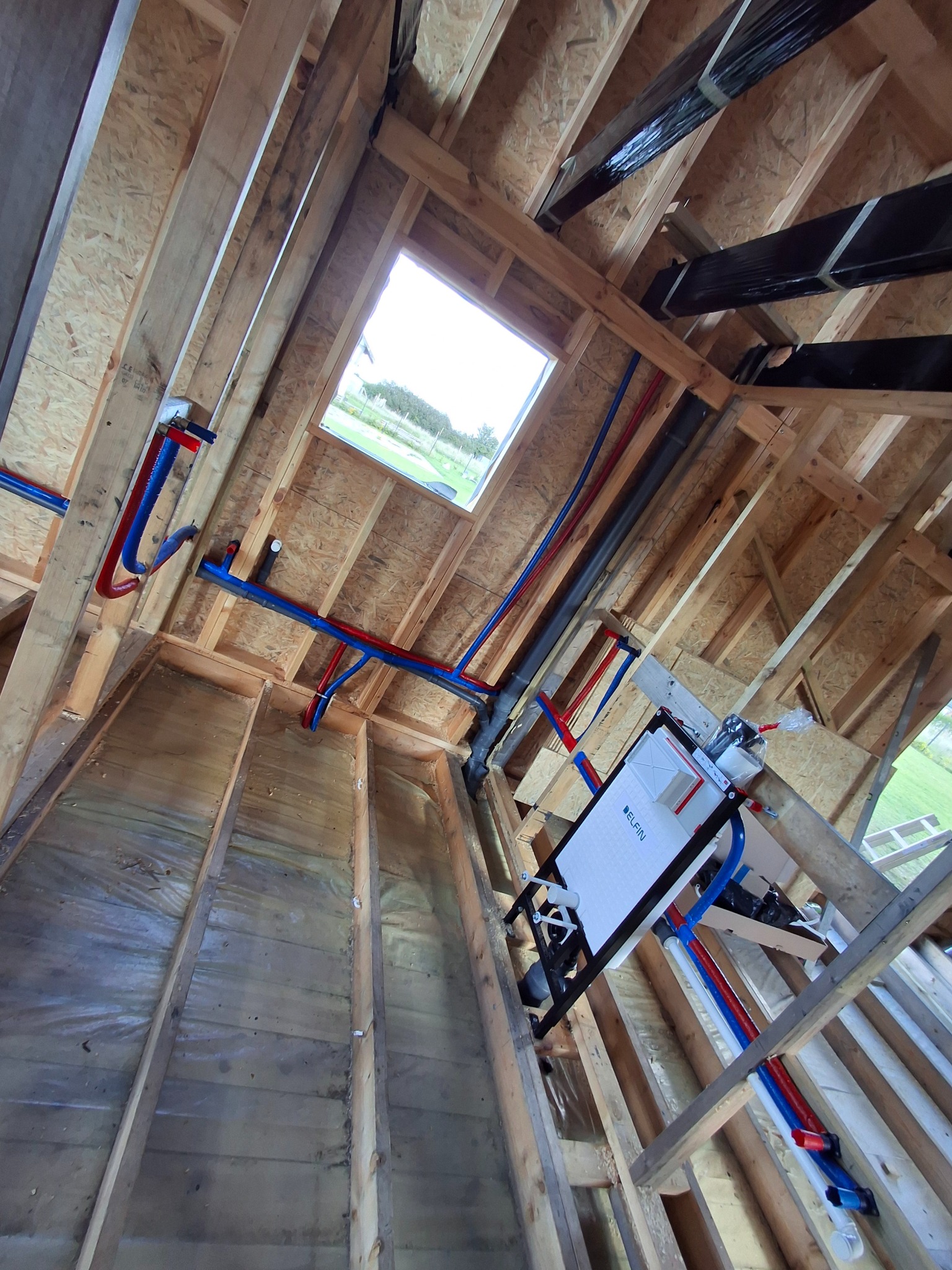 Instalacja wodno-kanalizacyjna w surowym wnętrzu poddasza: rury PEX w kolorach niebieskim i czerwonym, stelaż podtynkowy WC marki Elfin, widok z dołu na okno.