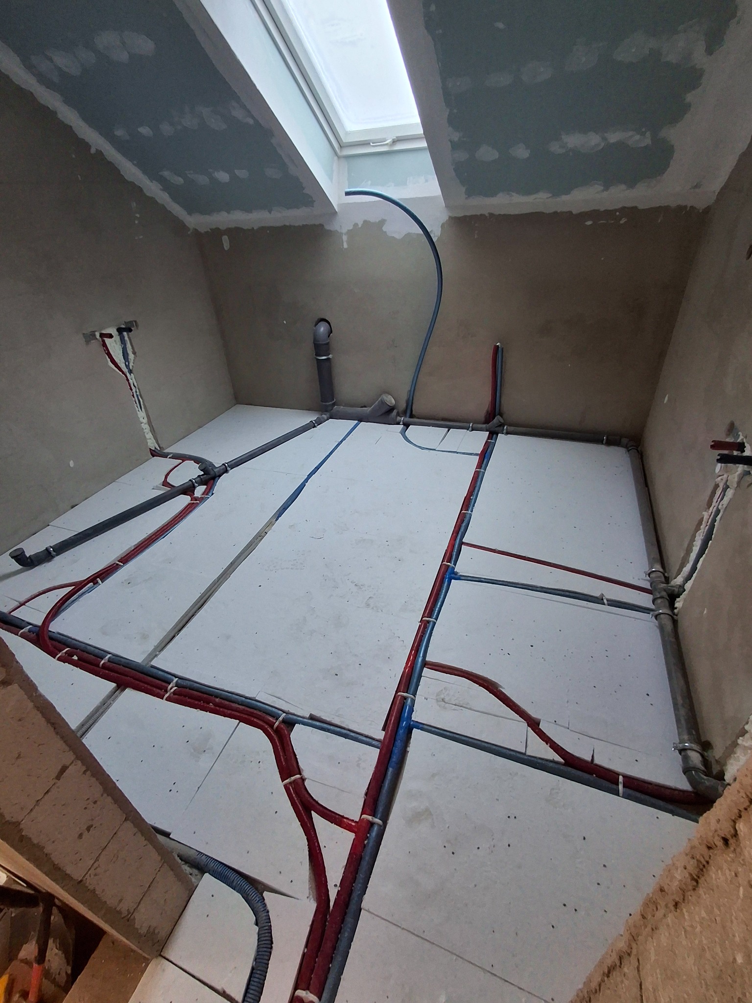 Instalacja rur grzewczych i wodnych na poddaszu z oknem dachowym, widoczne rury w kolorze czerwonym i niebieskim, przymocowane do białych płyt izolacyjnych.