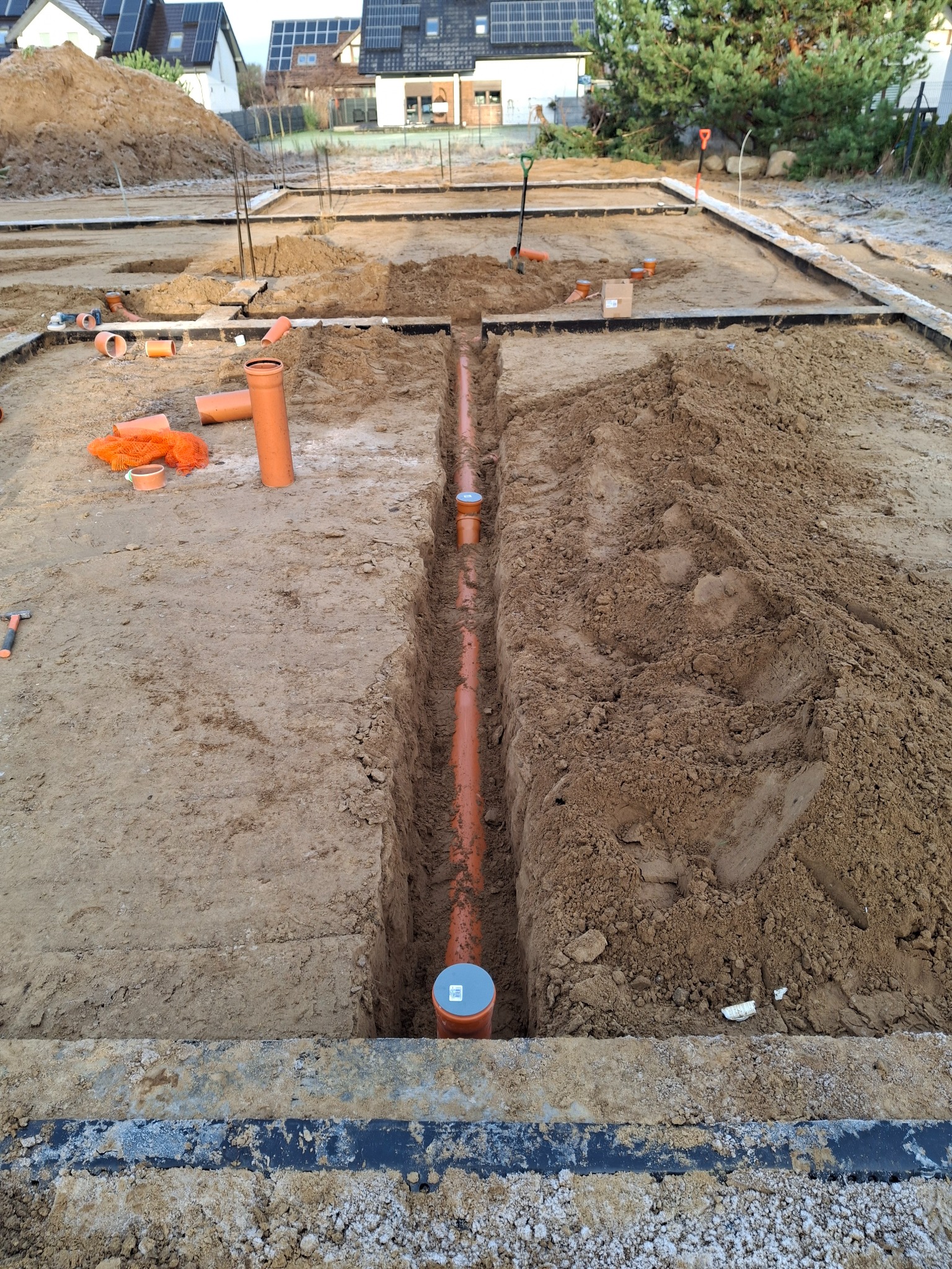 Wykop z ułożonymi rurami kanalizacyjnymi w ziemi, widoczne narzędzia i elementy konstrukcyjne. Prace ziemne w toku, przygotowanie do dalszych etapów budowy.