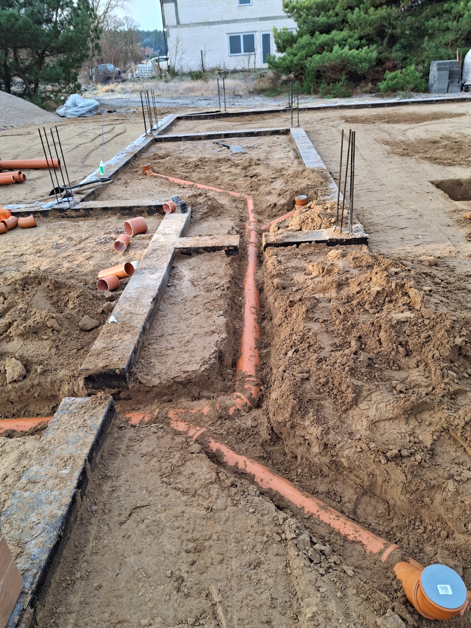 Pomarańczowe rury kanalizacyjne ułożone w wykopach fundamentowych na placu budowy domu jednorodzinnego. Widoczne zbrojenia i elementy szalunków.