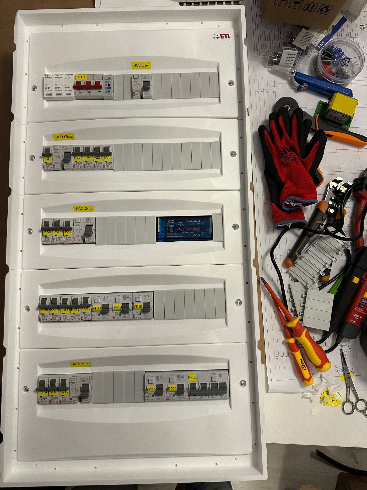 Rozdzielnica elektryczna z bezpiecznikami i wyłącznikami, widok z góry, obok narzędzia i schematy.  Elementy opisane etykietami.