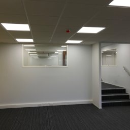 Paweł Rusek Malpl malowanie i remonty - Wnętrze biurowe po malowaniu: szara ściana z żółtym akcentem, okno wewnętrzne, wykładzina dywanowa w paski, schody z poręczą. Widoczne gniazdka elektryczne przy podłodze.