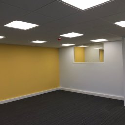 Paweł Rusek Malpl malowanie i remonty - Pomalowane biuro: Żółta ściana kontrastuje z białą, szara podłoga i sufit z kwadratowymi lampami. Widoczne gniazdka elektryczne i okno w ścianie.