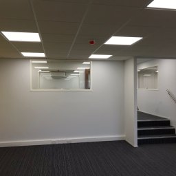 Paweł Rusek Malpl malowanie i remonty - Jasne biuro po remoncie: świeżo pomalowane ściany, nowe panele sufitowe z wbudowanym oświetleniem, wykładzina dywanowa w paski i schody z metalową barierką.