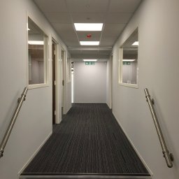 Paweł Rusek Malpl malowanie i remonty - Wnętrze biurowego korytarza z szarymi ścianami, wykładziną w paski i metalowymi poręczami. Widoczne okna i drzwi prowadzące do pomieszczeń. Oświetlenie sufitowe.