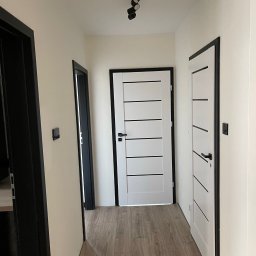 Paweł Rusek Malpl malowanie i remonty - Jasny korytarz po remoncie z trzema parami drzwi w czarnych futrynach i drewnianą podłogą. Na suficie czarna lampa z trzema reflektorami.