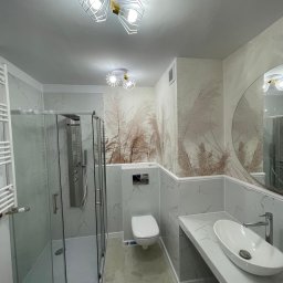 Paweł Rusek Malpl malowanie i remonty - Nowoczesna łazienka z kabiną prysznicową, umywalką nablatową i toaletą podwieszaną. Na ścianach płytki imitujące marmur i tapeta z motywem roślinnym. Oświetlenie sufitowe.