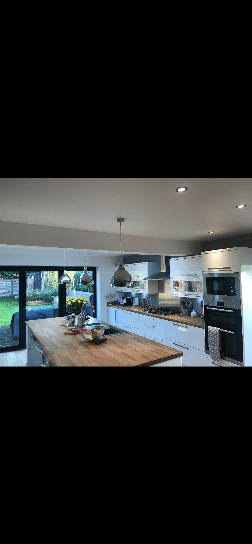 Nowoczesna kuchnia z wyspą i widokiem na ogród przez przesuwne drzwi. Białe szafki, drewniany blat, metalowe lampy wiszące. Jasne, przestronne wnętrze.