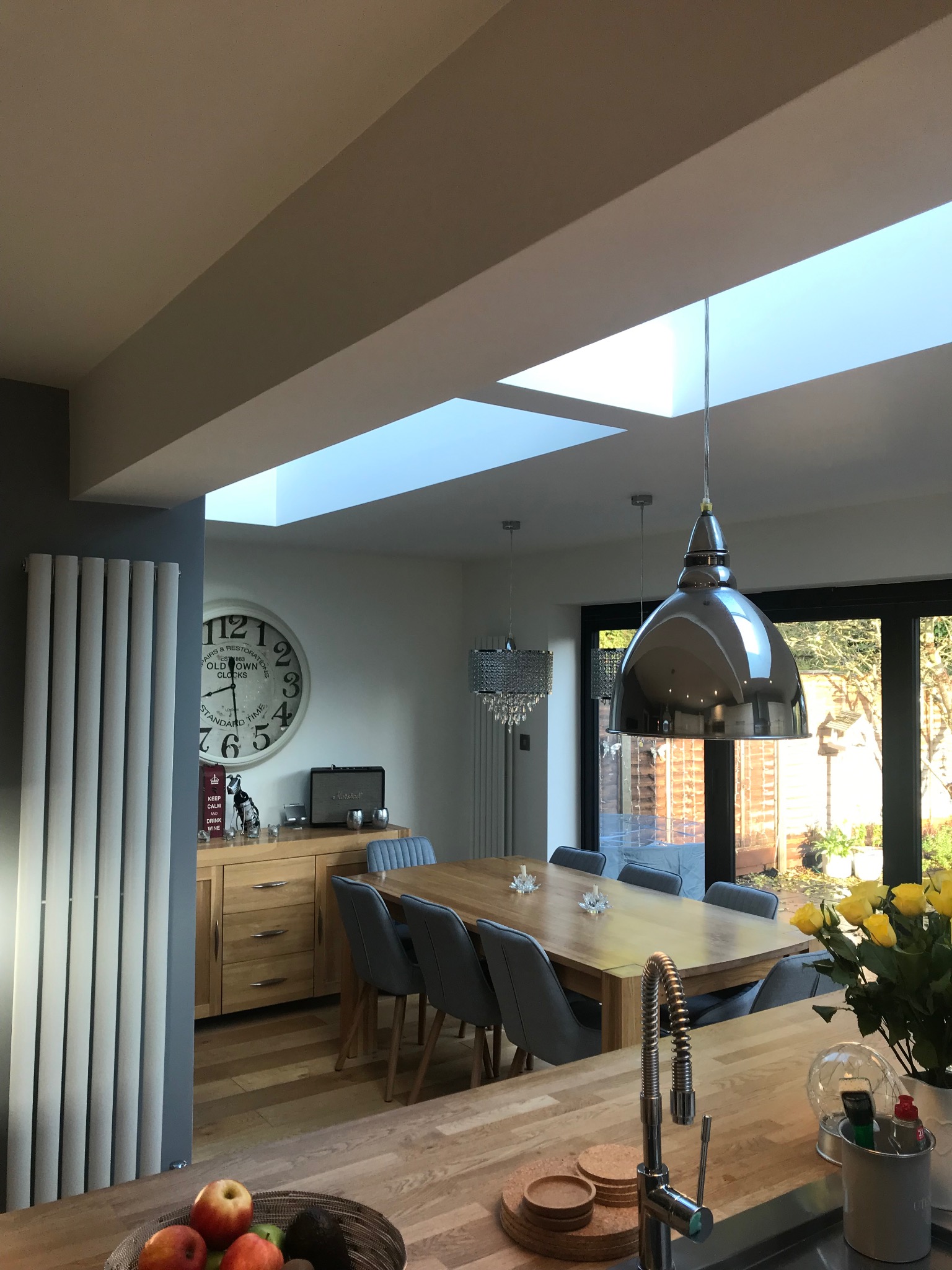 Jasne wnętrze jadalni z drewnianym stołem, szarymi krzesłami i nowoczesnym oświetleniem. Okno dachowe wpuszcza naturalne światło. Widoczny grzejnik i dekoracje.