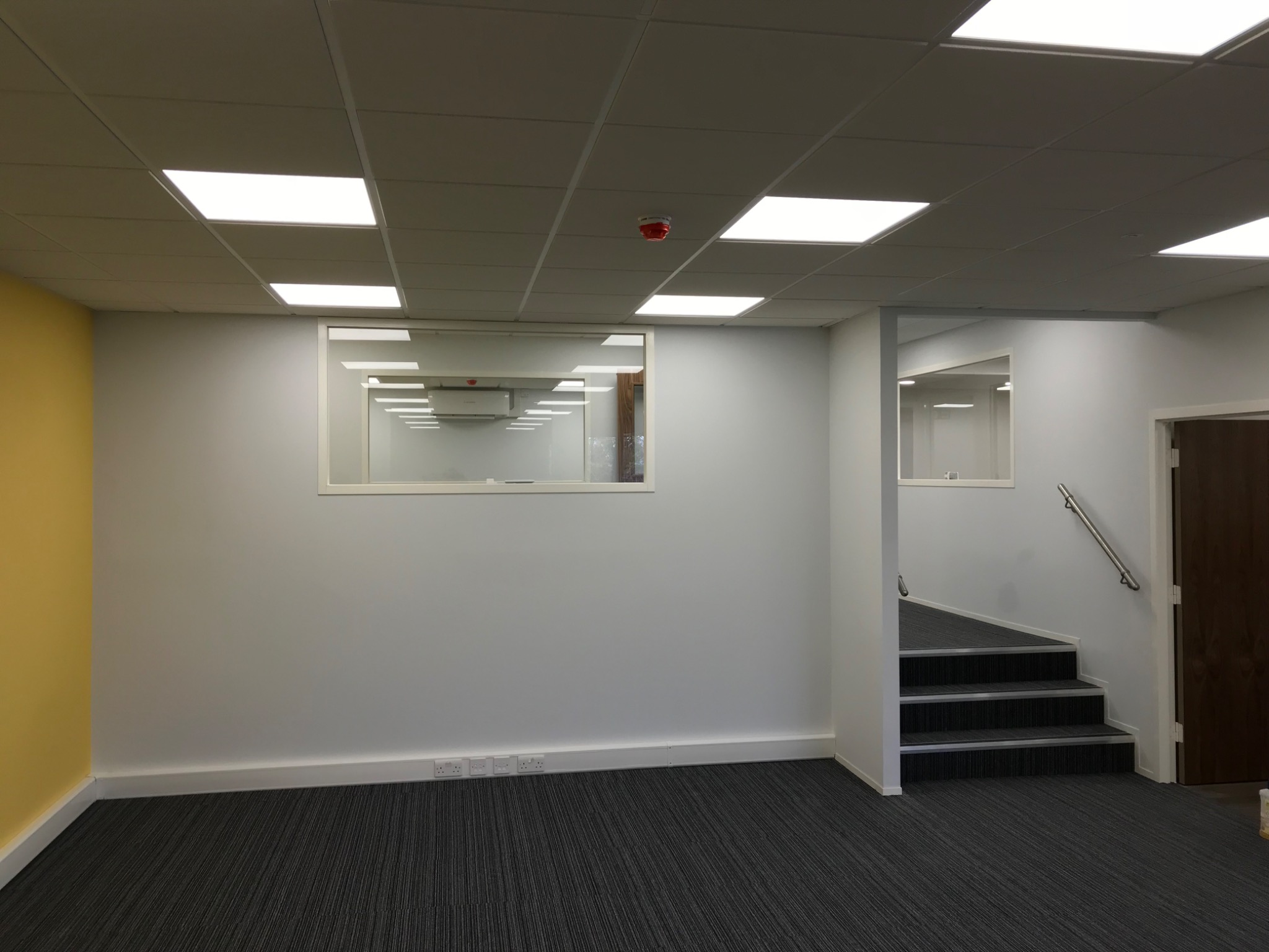 Wnętrze biurowe po malowaniu: szara ściana z żółtym akcentem, okno wewnętrzne, wykładzina dywanowa w paski, schody z poręczą. Widoczne gniazdka elektryczne przy podłodze.