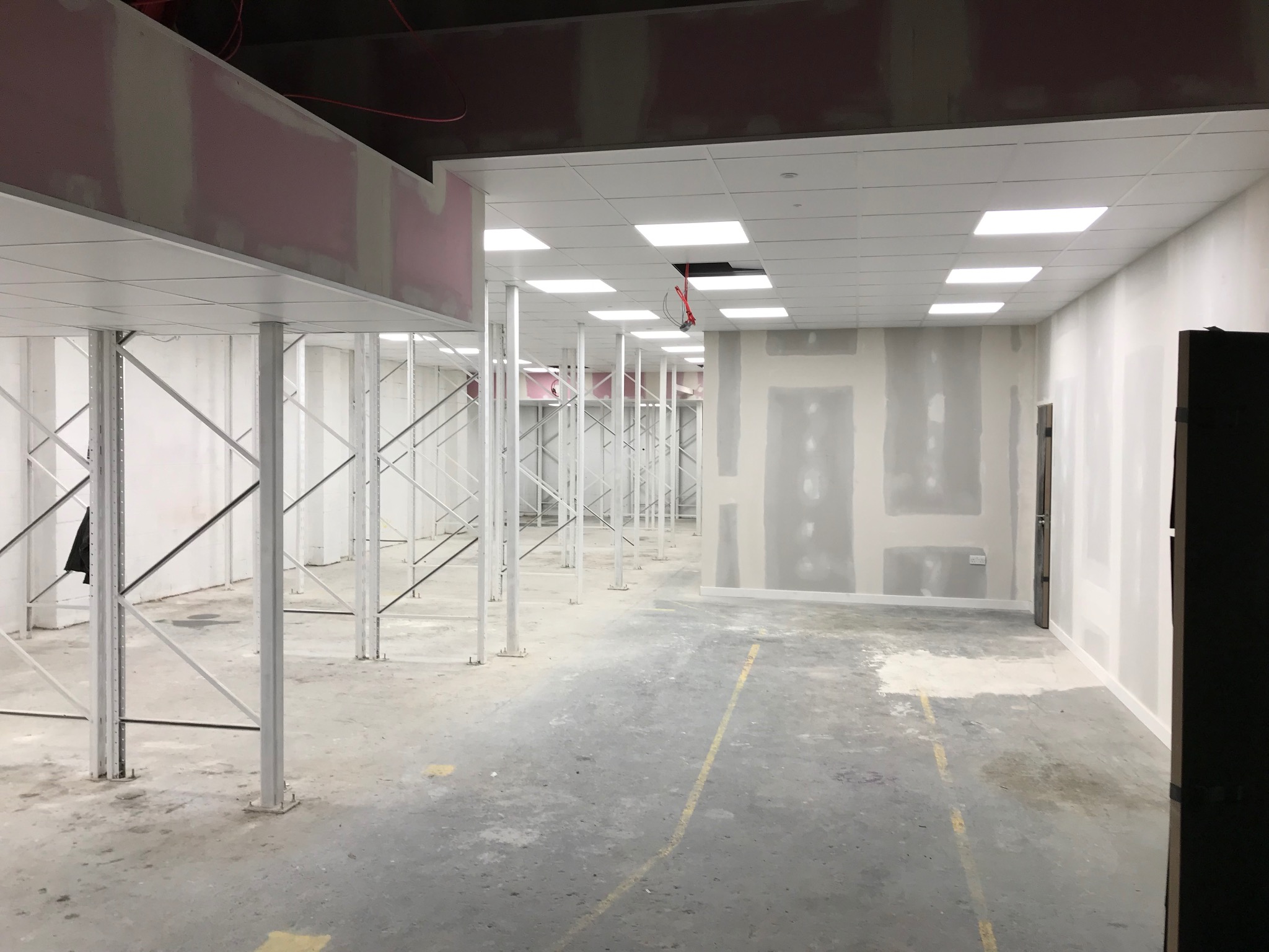 Wnętrze komercyjne w trakcie prac wykończeniowych: szpachlowane ściany, sufit podwieszany z oświetleniem, metalowe podpory konstrukcyjne. Widok na przestrzeń w trakcie przygotowania.