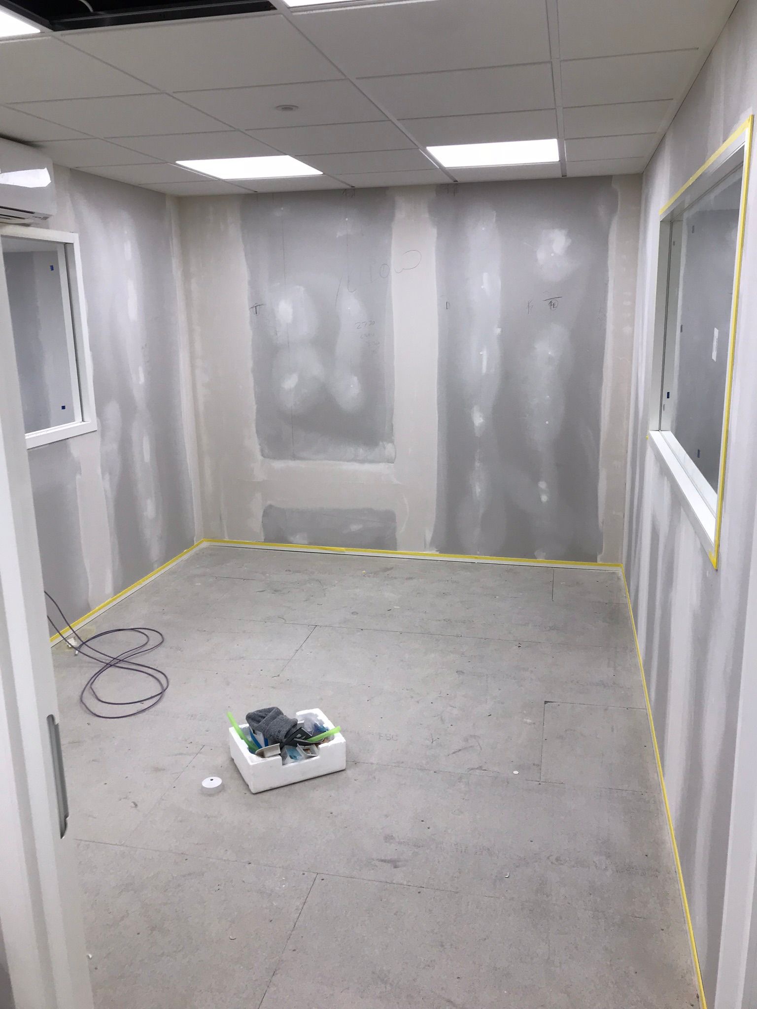 Pokój w trakcie szpachlowania ścian, zabezpieczony taśmą malarską przy podłodze i oknach. Na podłodze leżą narzędzia w białym pojemniku i zwinięty kabel.