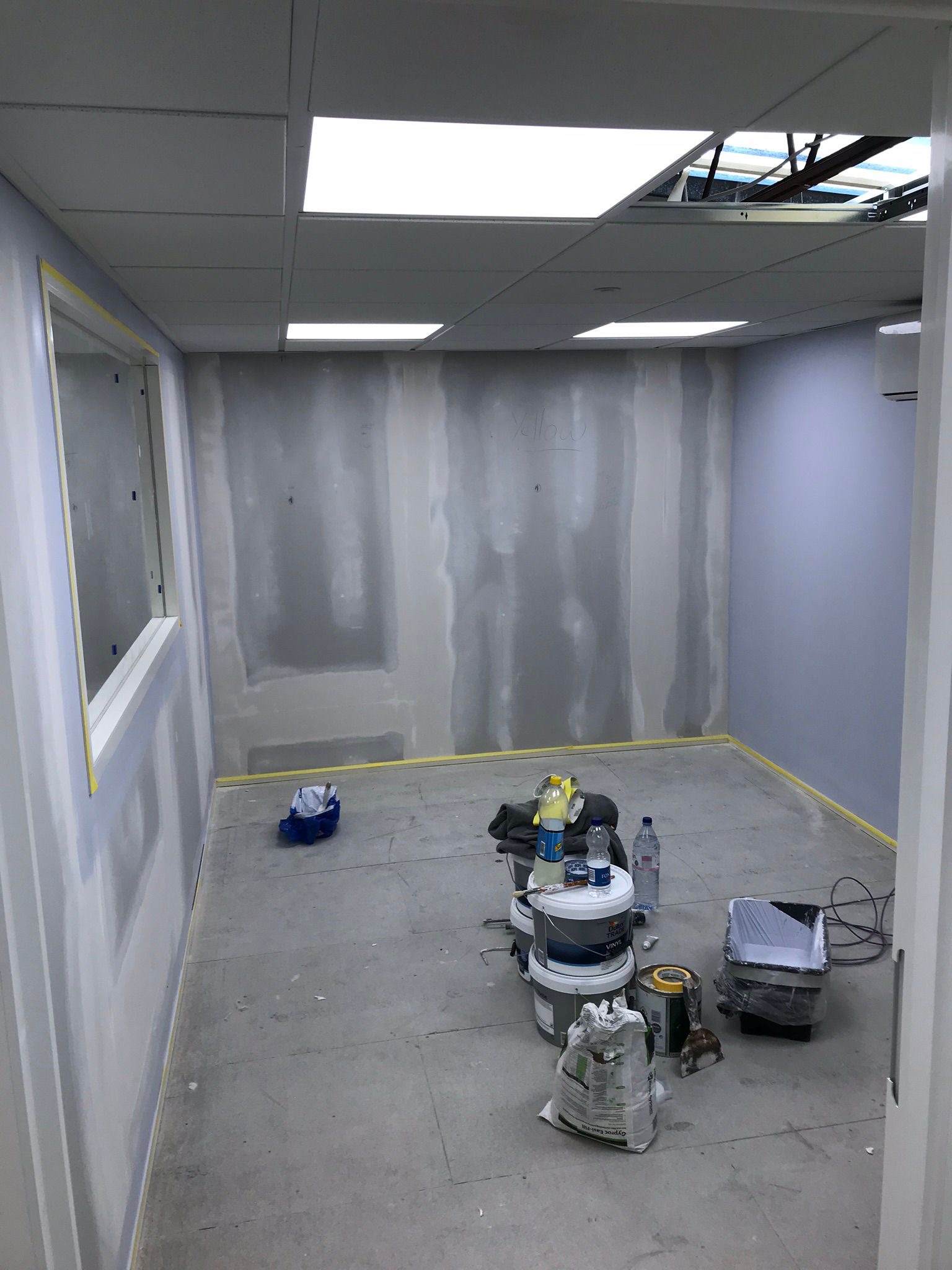 Wnętrze biura w trakcie szpachlowania: ściany częściowo pokryte gładzią, zabezpieczone taśmą, widoczne narzędzia i materiały budowlane, sufit podwieszany z oświetleniem.