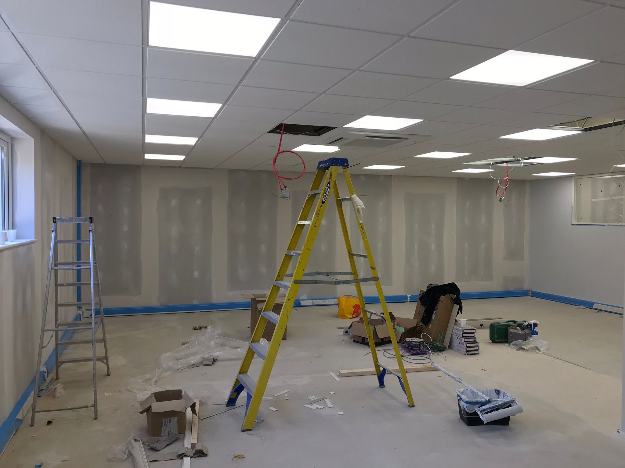 Wnętrze biura w trakcie szpachlowania ścian, widoczne drabiny, sufit podwieszany z oświetleniem, zabezpieczone podłogi i ściany. Prace wykończeniowe w toku.