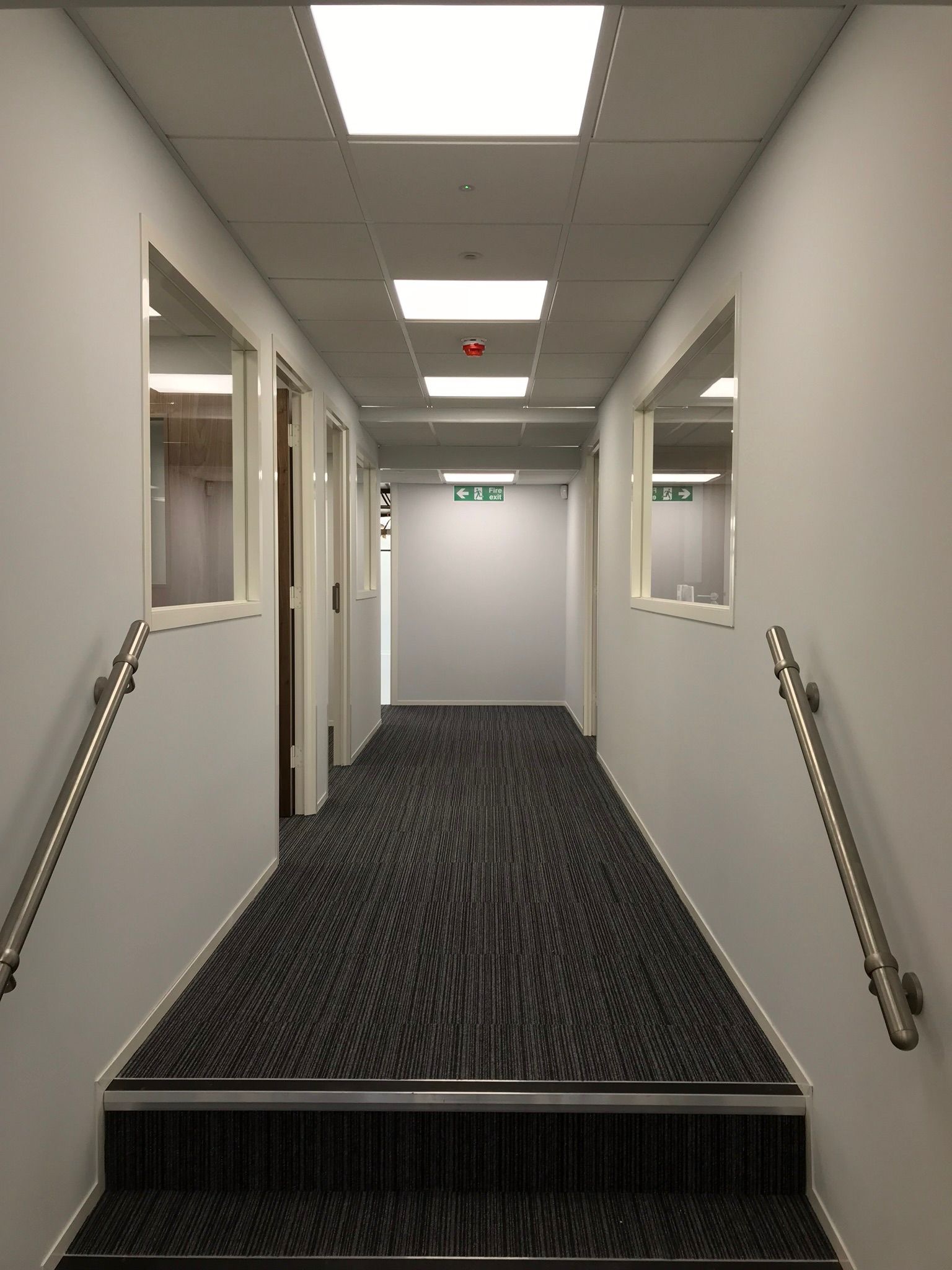 Wnętrze biurowego korytarza z szarymi ścianami, wykładziną w paski i metalowymi poręczami. Widoczne okna i drzwi prowadzące do pomieszczeń. Oświetlenie sufitowe.