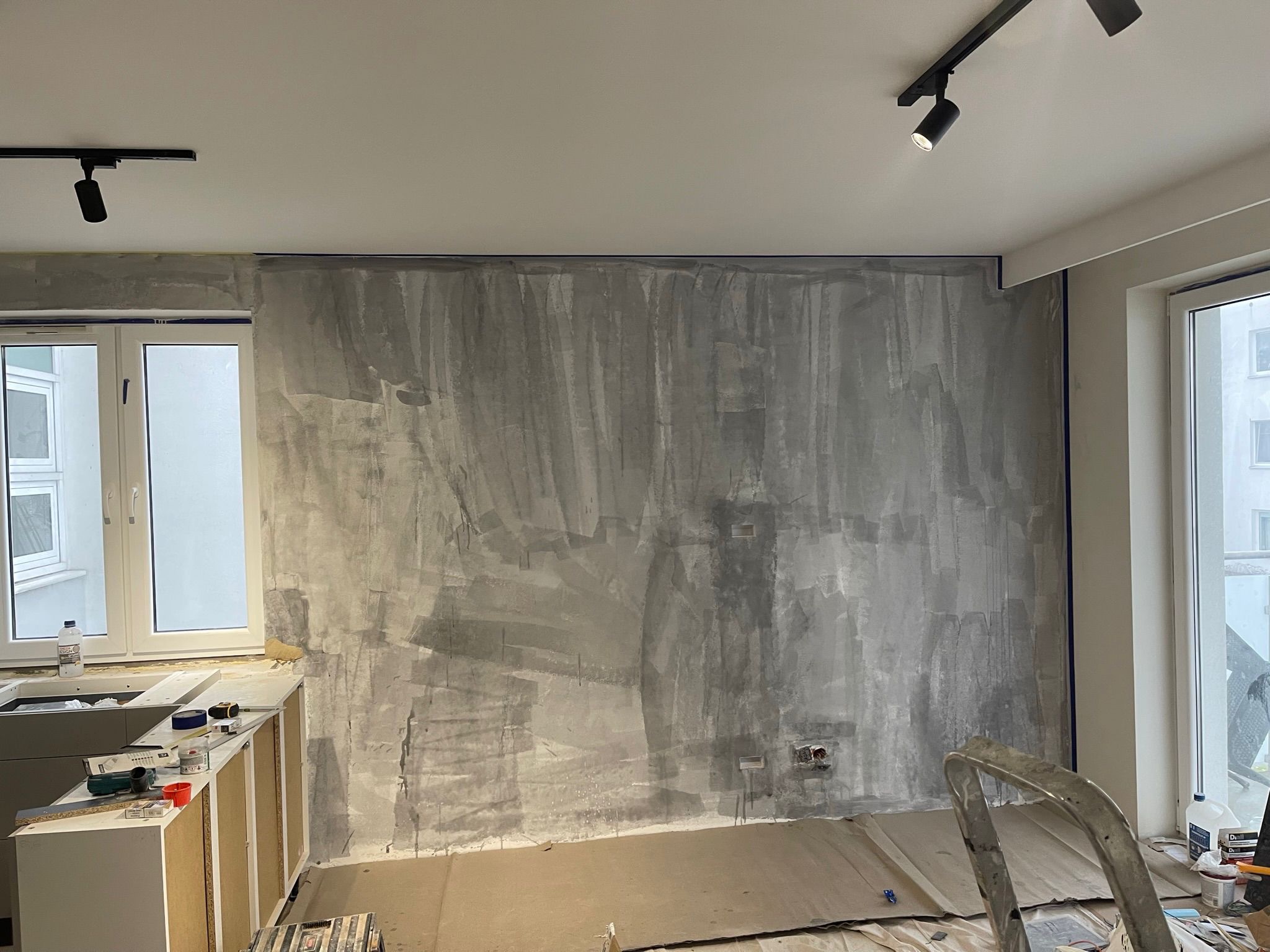 Ściana po szpachlowaniu, przygotowana do malowania. Widoczne ślady po szpachli, taśma malarska przy oknach i suficie. Na podłodze kartony zabezpieczające.