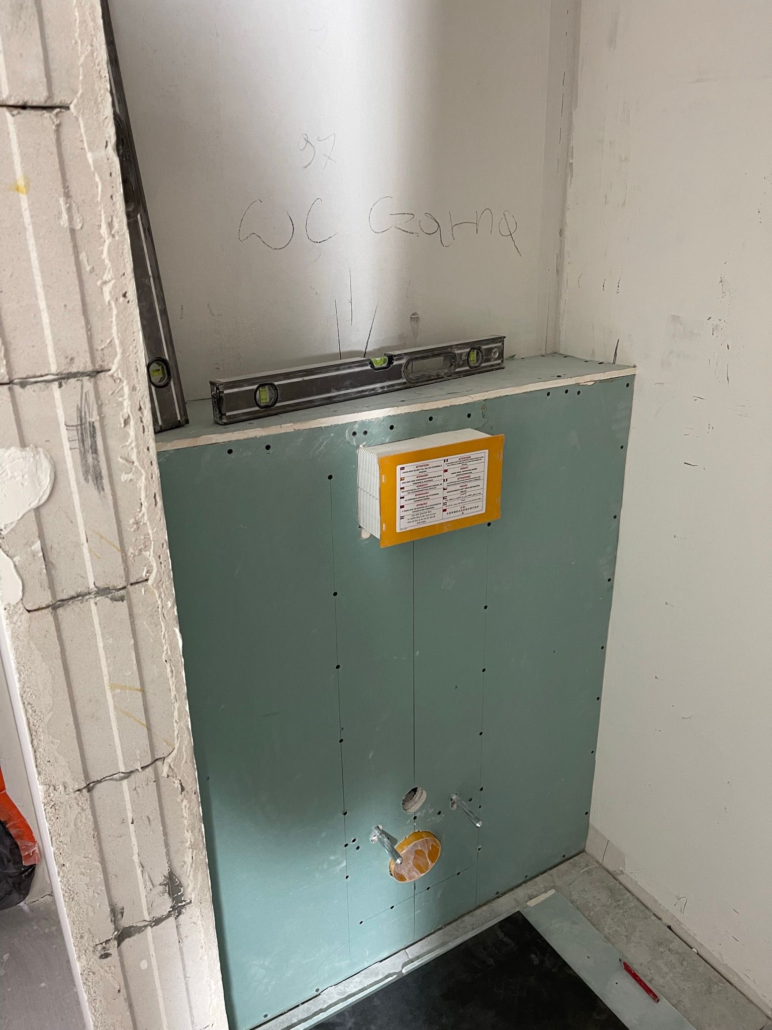 Montaż płyty gipsowej w łazience, widoczne poziomice, instalacja wodna i oznaczenia na ścianie. Prace wykończeniowe w toalecie, przygotowanie do dalszych etapów remontu.