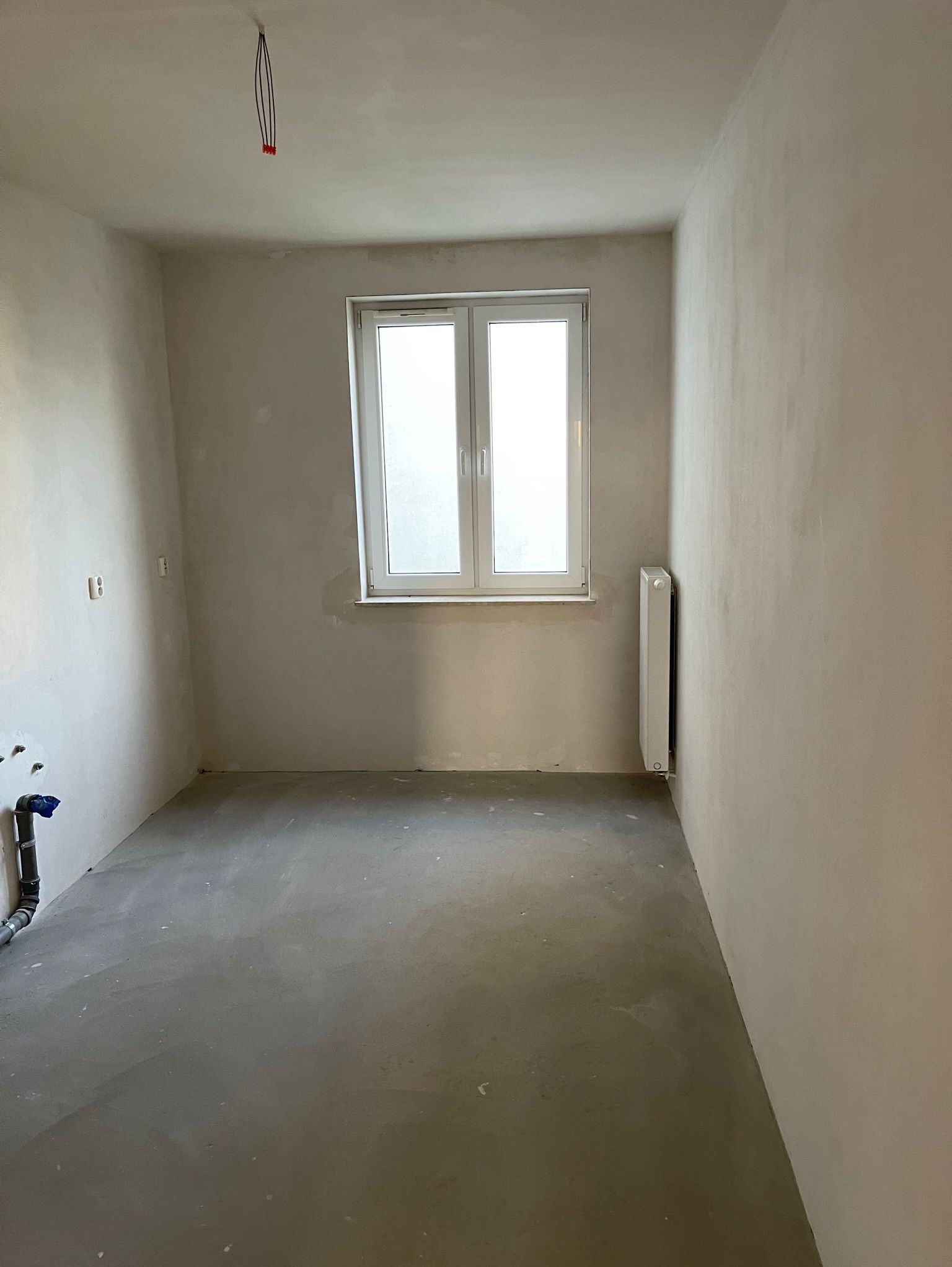 Wnętrze pokoju po szpachlowaniu ścian, z widocznym oknem, grzejnikiem i instalacją hydrauliczną. Przygotowanie do dalszych prac wykończeniowych.
