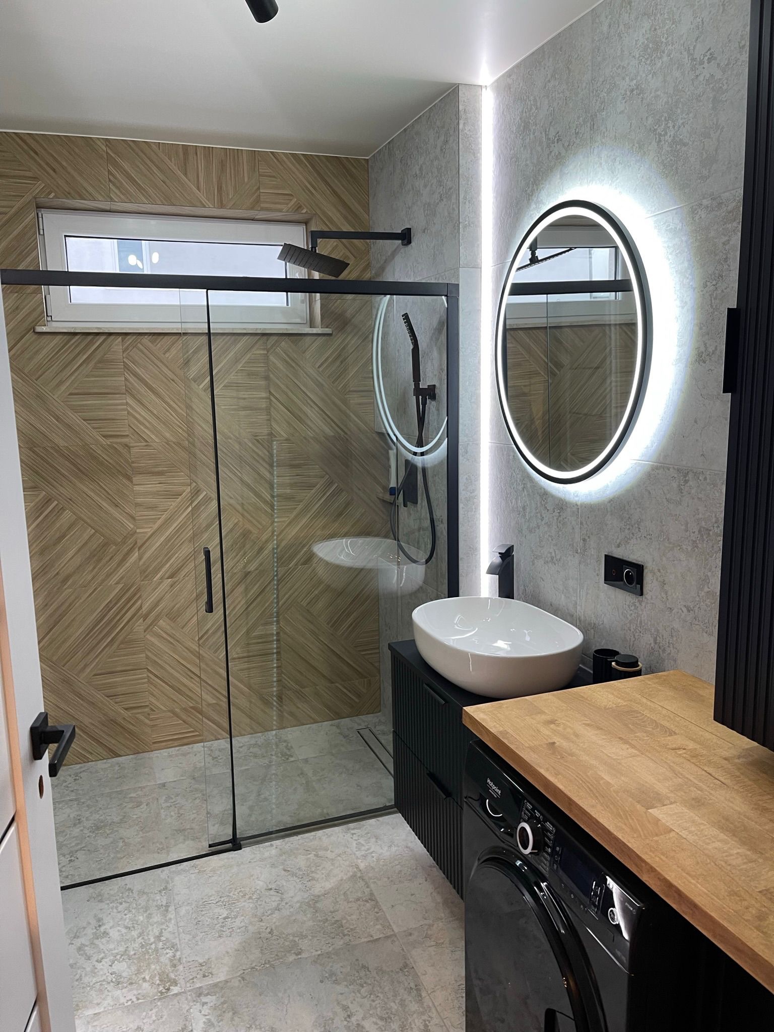 Nowoczesna łazienka z kabiną prysznicową typu walk-in, umywalką nablatową i okrągłym lustrem z podświetleniem LED. Uwagę zwraca drewnopodobna okładzina w strefie prysznica.