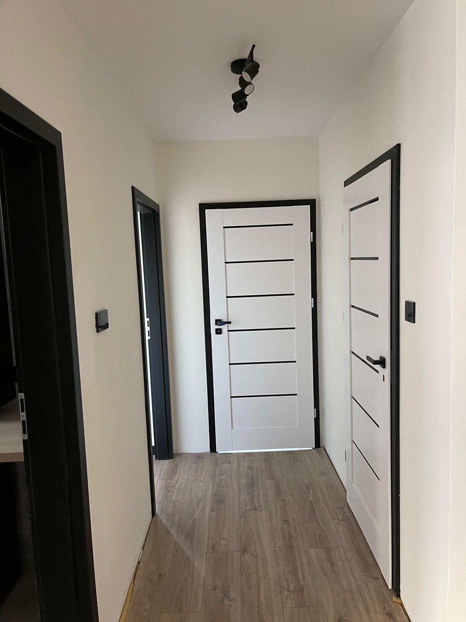 Jasny korytarz po remoncie z trzema parami drzwi w czarnych futrynach i drewnianą podłogą. Na suficie czarna lampa z trzema reflektorami.