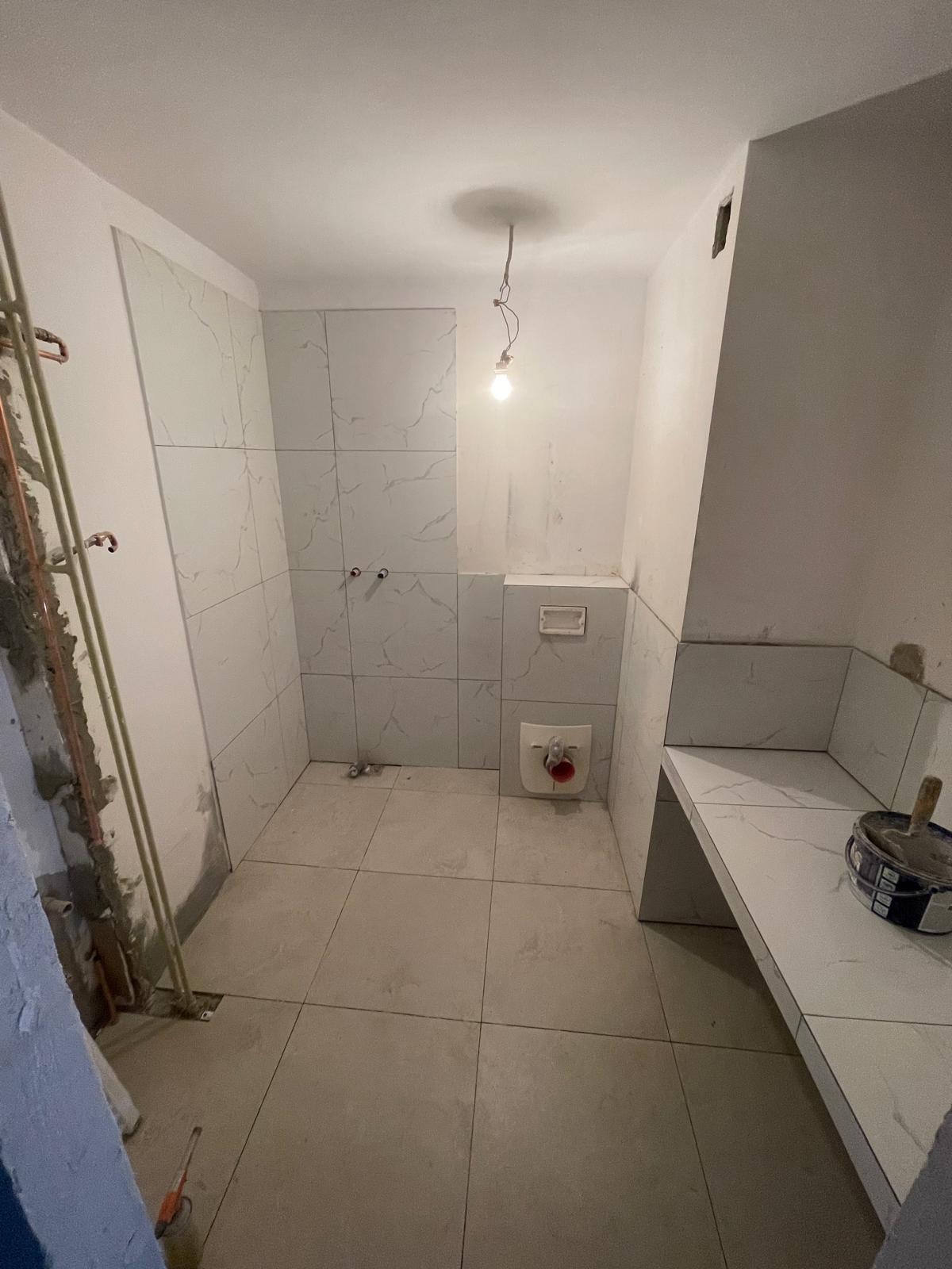 Remont łazienki w trakcie: białe płytki imitujące marmur na ścianach i podłodze, częściowo zamontowana toaleta podwieszana, widoczne rury i puszka z farbą.