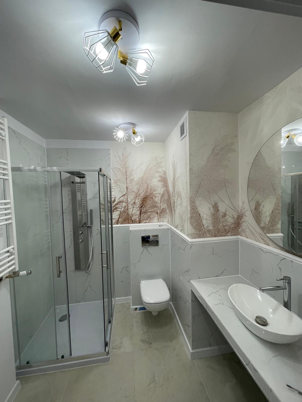 Nowoczesna łazienka z kabiną prysznicową, umywalką nablatową i toaletą podwieszaną. Na ścianach płytki imitujące marmur i tapeta z motywem roślinnym. Oświetlenie sufitowe.