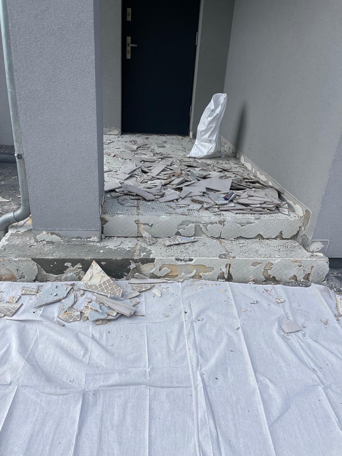 Demontaż płytek na schodach wejściowych do budynku. Widoczne resztki kleju i gruz. Prace przygotowawcze do położenia nowej glazury. Zabezpieczenie podłoża folią malarską.