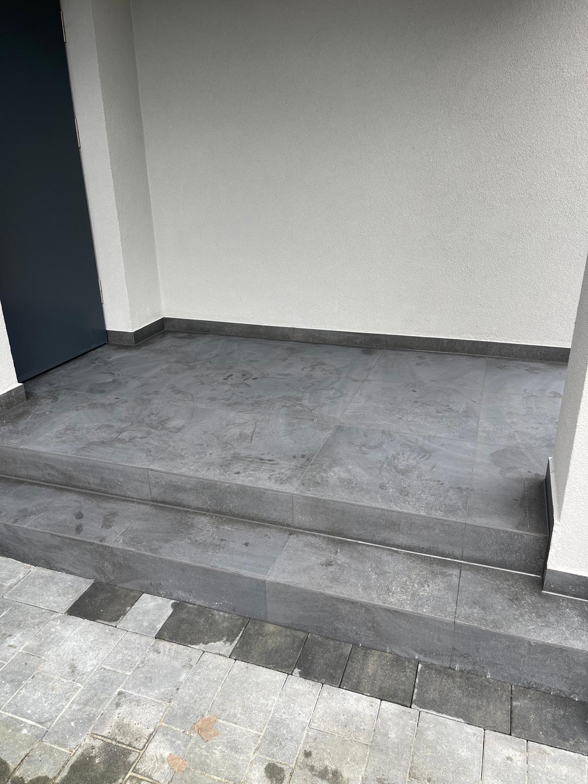 Wykończone płytkami schody wejściowe w odcieniach szarości, z widoczną fakturą kamienia. Minimalistyczny design, jasna elewacja budynku i ciemne drzwi wejściowe.