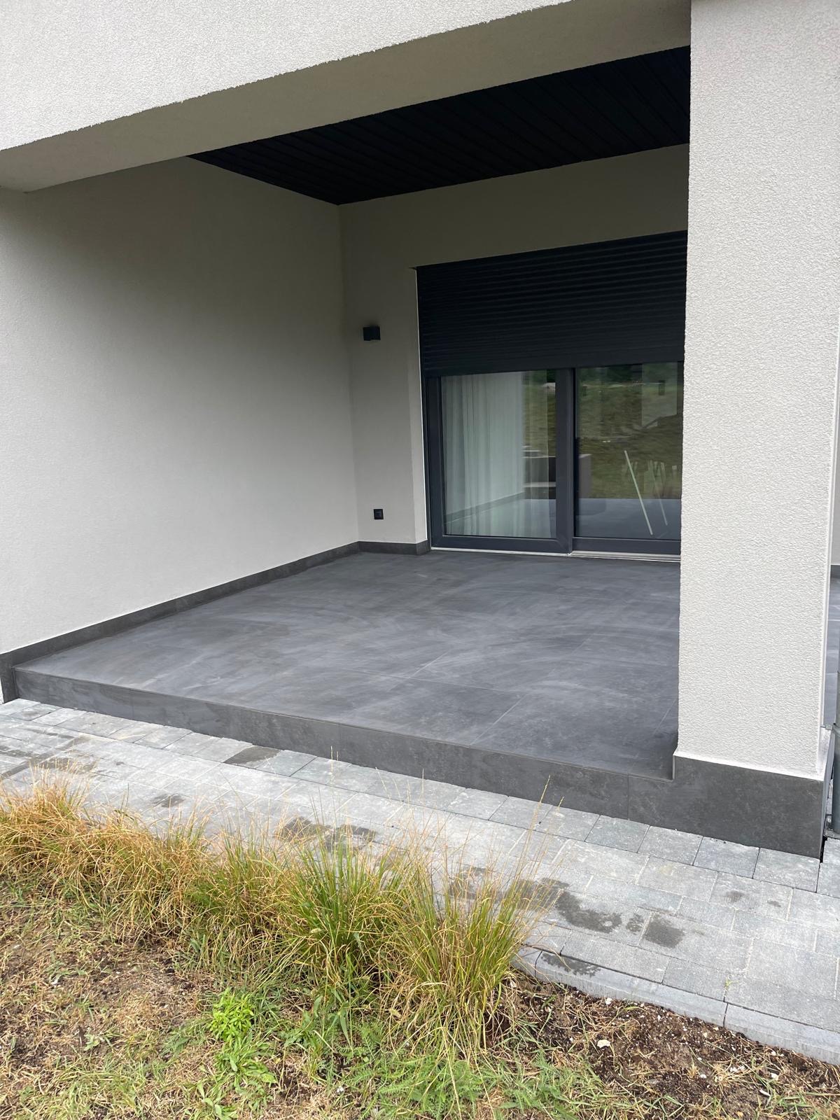 Zadaszony taras z szarą, gładką podłogą z płytek, widok z zewnątrz. Ściany w jasnym odcieniu szarości, czarne rolety okienne. Minimalistyczny design.