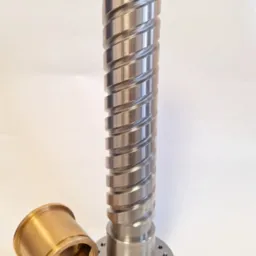 Metalowy wał śrubowy z gwintem trapezowym i kołnierzem, obok tuleja z brązu. Precyzyjna obróbka skrawaniem, gładka powierzchnia, widoczne ślady obróbki.