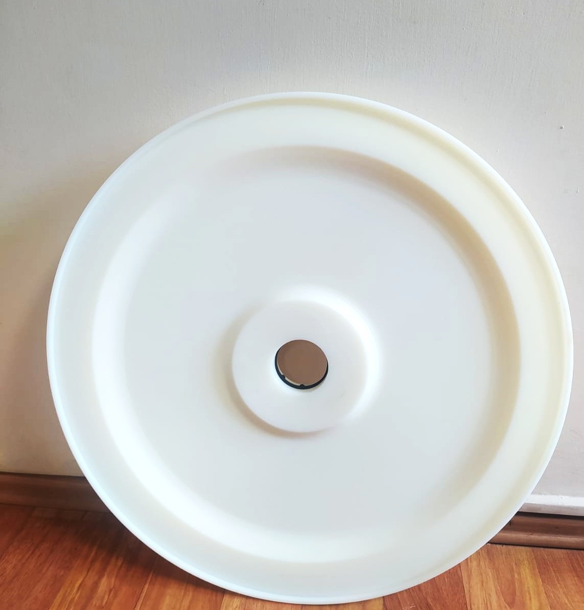 Biały, okrągły element z tworzywa z centralnym otworem, leżący na drewnianej podłodze przy ścianie. Gładka powierzchnia, symetryczny kształt.