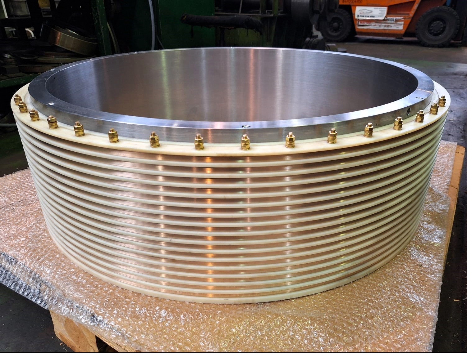 Precyzyjnie wykonany, duży, cylindryczny element z metalu i tworzywa, z licznymi żebrami i śrubami, leżący na palecie. Widoczny wózek widłowy w tle.