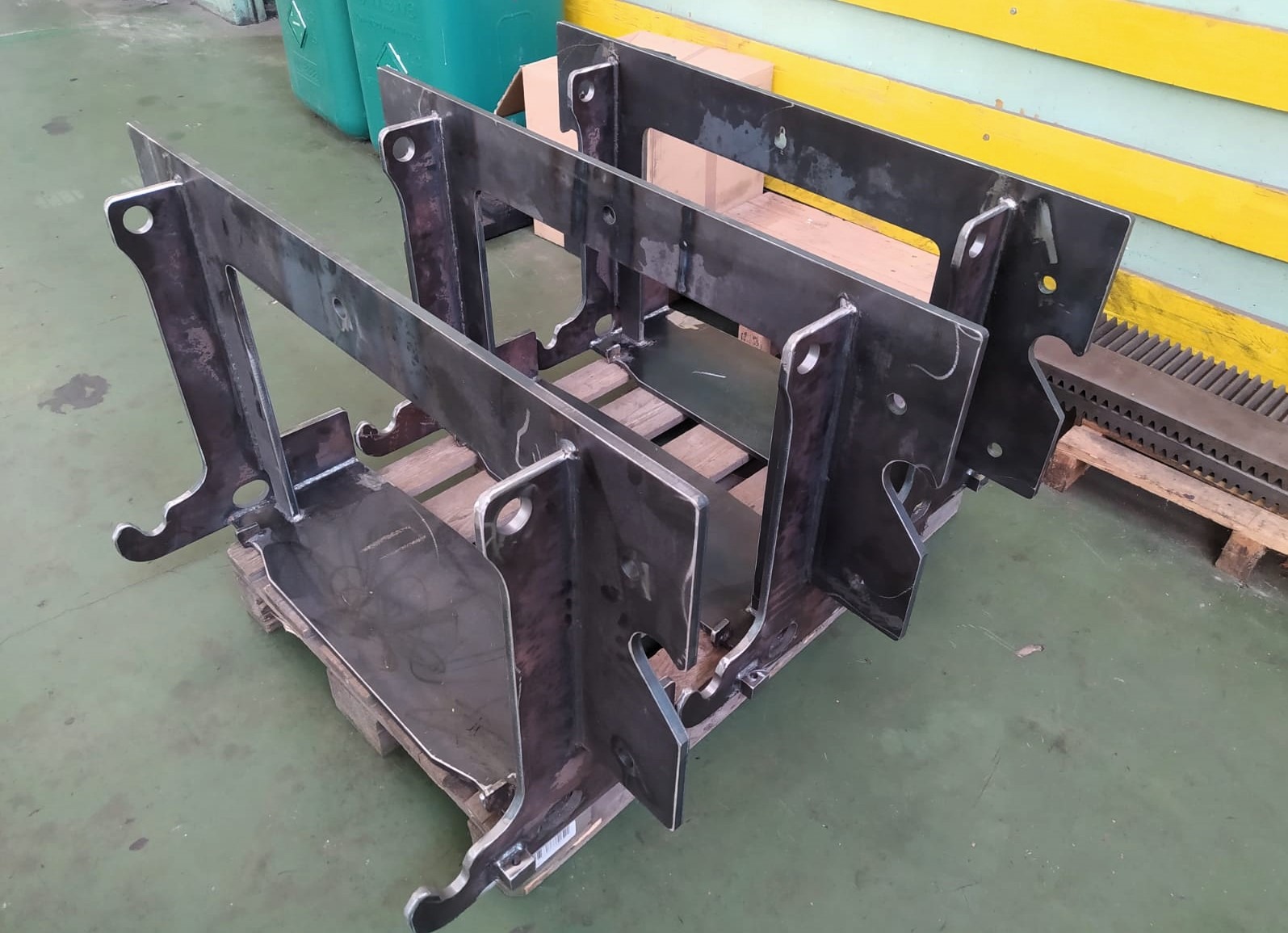 Surowe, metalowe elementy konstrukcyjne na palecie, po obróbce. Widoczne otwory i wycięcia. Tło stanowią elementy hali produkcyjnej i skrzynia.