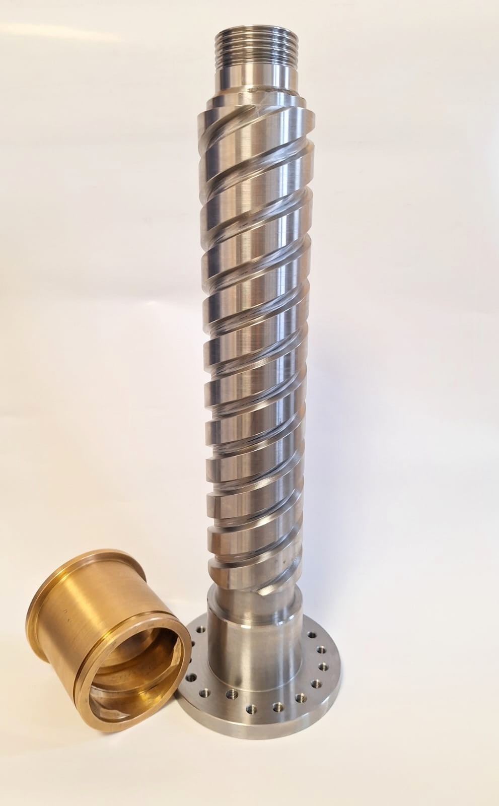 Metalowy wał śrubowy z gwintem trapezowym i kołnierzem, obok tuleja z brązu. Precyzyjna obróbka skrawaniem, gładka powierzchnia, widoczne ślady obróbki.