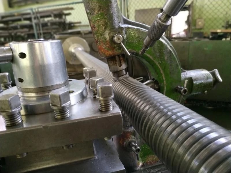 Metalowa śruba z gwintem trapezowym obrabiana na tokarce. Widoczne elementy tokarki, uchwyt i narzędzie skrawające. Detal z bliska, oświetlenie warsztatowe.